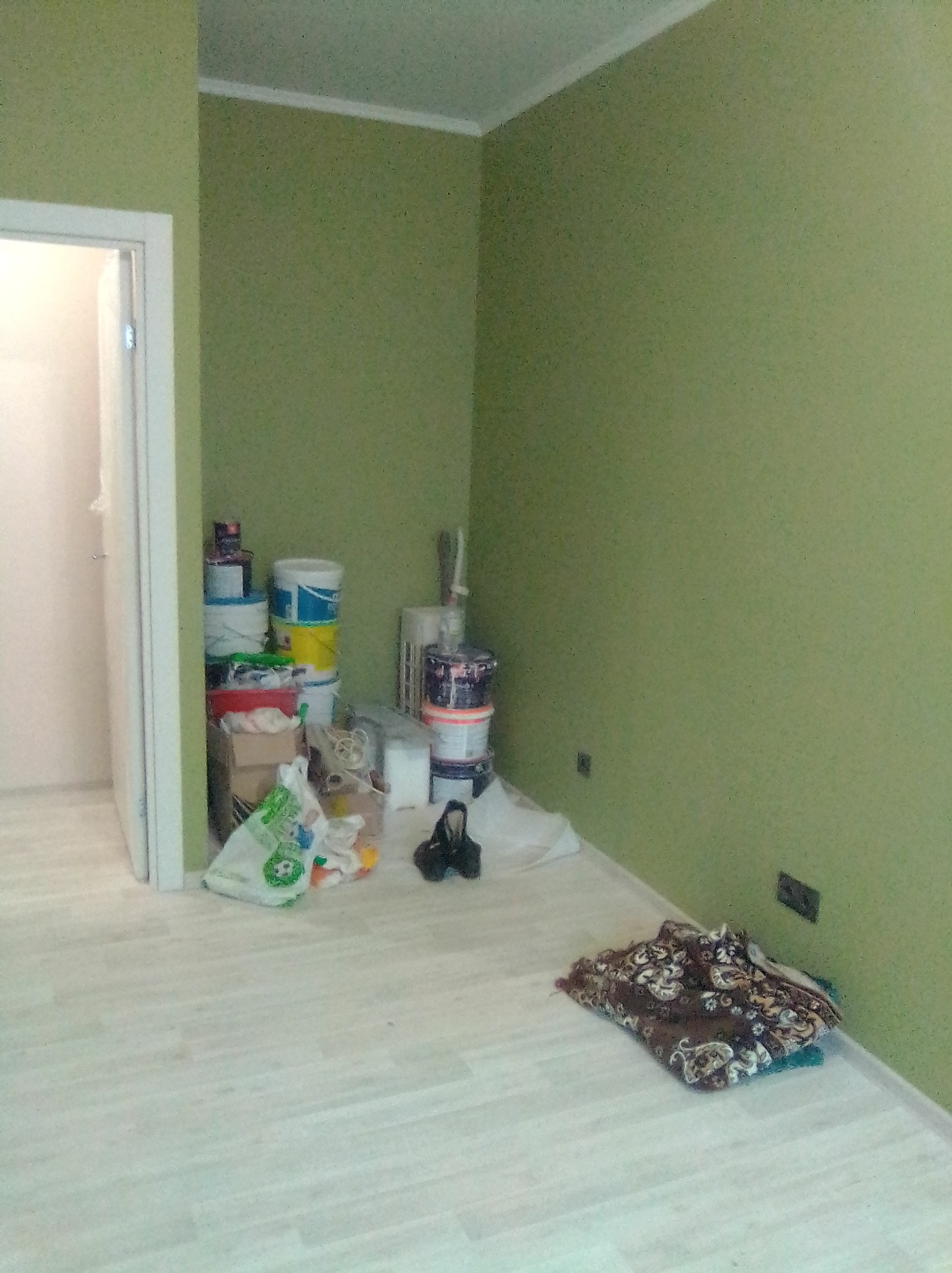 Pomalowany na zielono pokój z jasną podłogą. Przy ścianie stoją wiadra z farbą i inne akcesoria malarskie. Widoczny fragment drzwi.