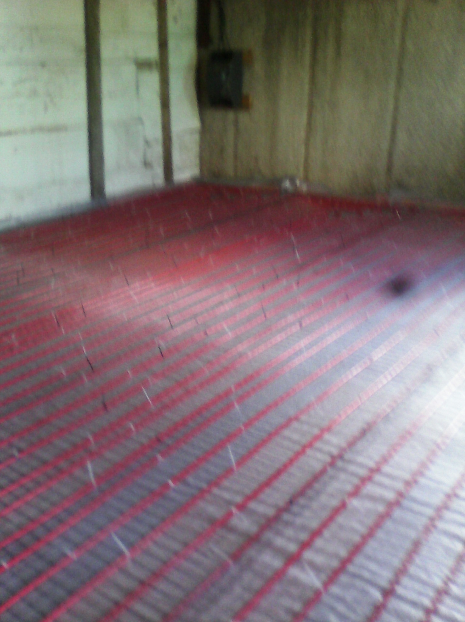 Czerwona instalacja ogrzewania podłogowego, ułożona równolegle na szarej powierzchni w surowym wnętrzu. Widoczne ściany w tle i skrzynka elektryczna.