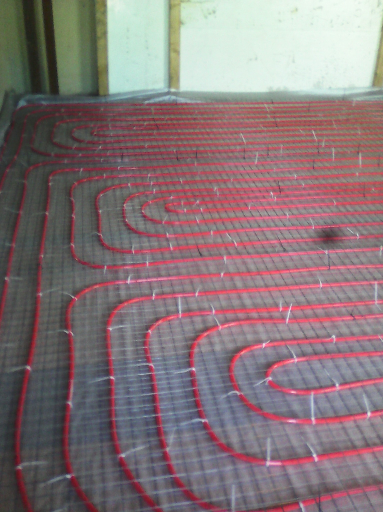 Czerwone rurki ogrzewania podłogowego ułożone na siatce, gotowe do zalania. Widoczne białe mocowania i fragment ściany w tle.