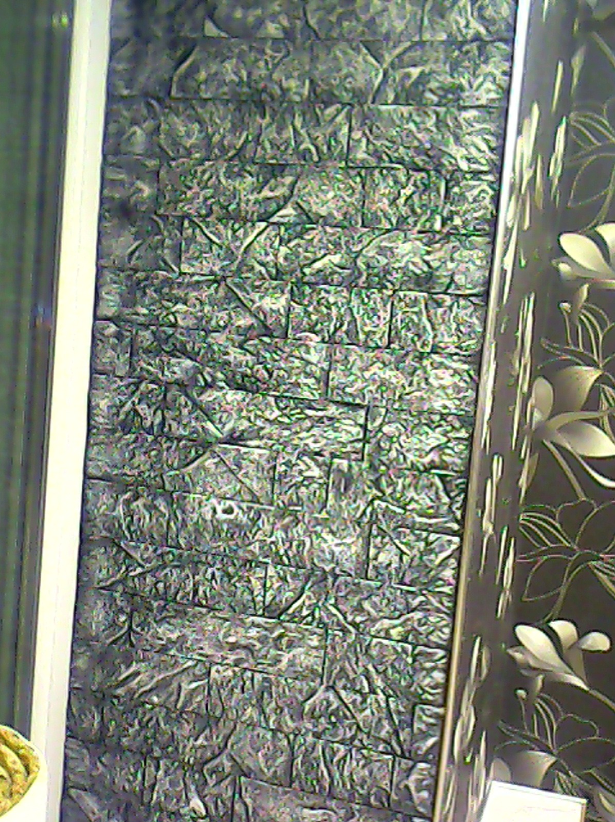 Szara, teksturowana ściana z efektem 3D, ułożona z małych, nieregularnych płytek. Obok widoczny fragment ściany z ciemną tapetą w kwiaty i biała listwa.