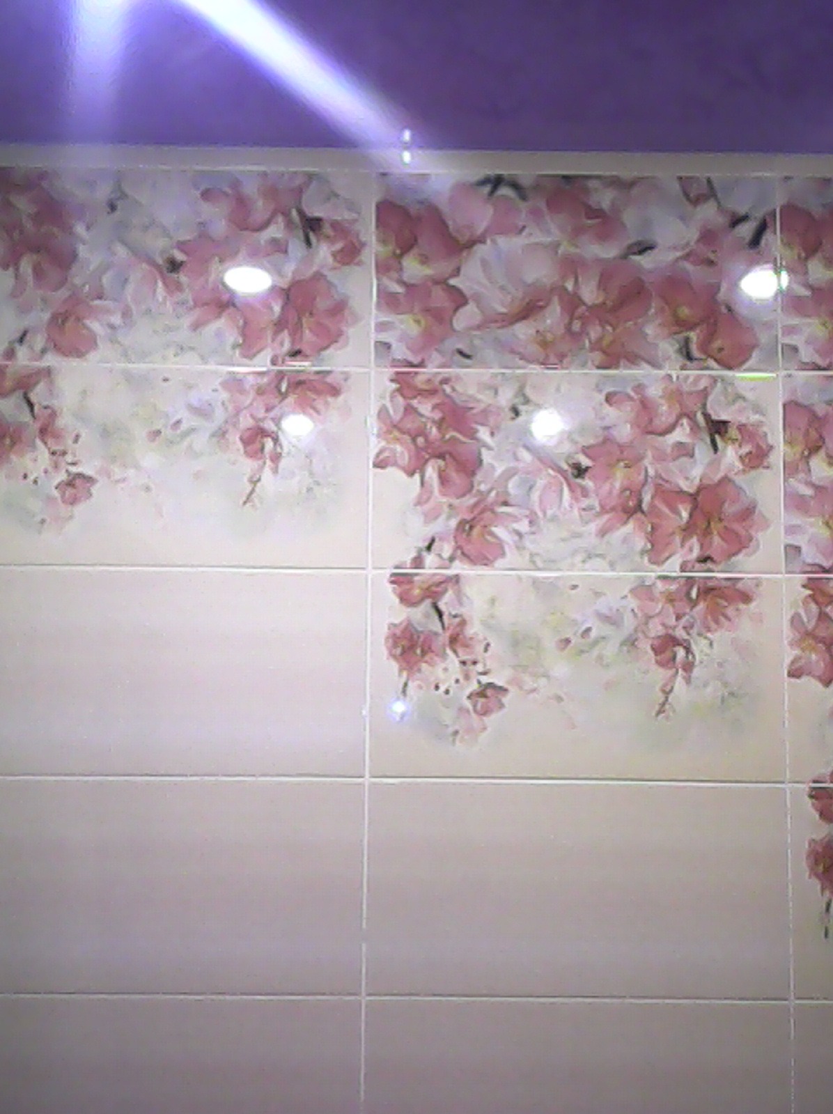 Ściana wyłożona jasnymi płytkami z motywem kwiatowym, z widocznym fragmentem fioletowego sufitu. Delikatny wzór kwiatów dodaje wnętrzu subtelnego uroku.