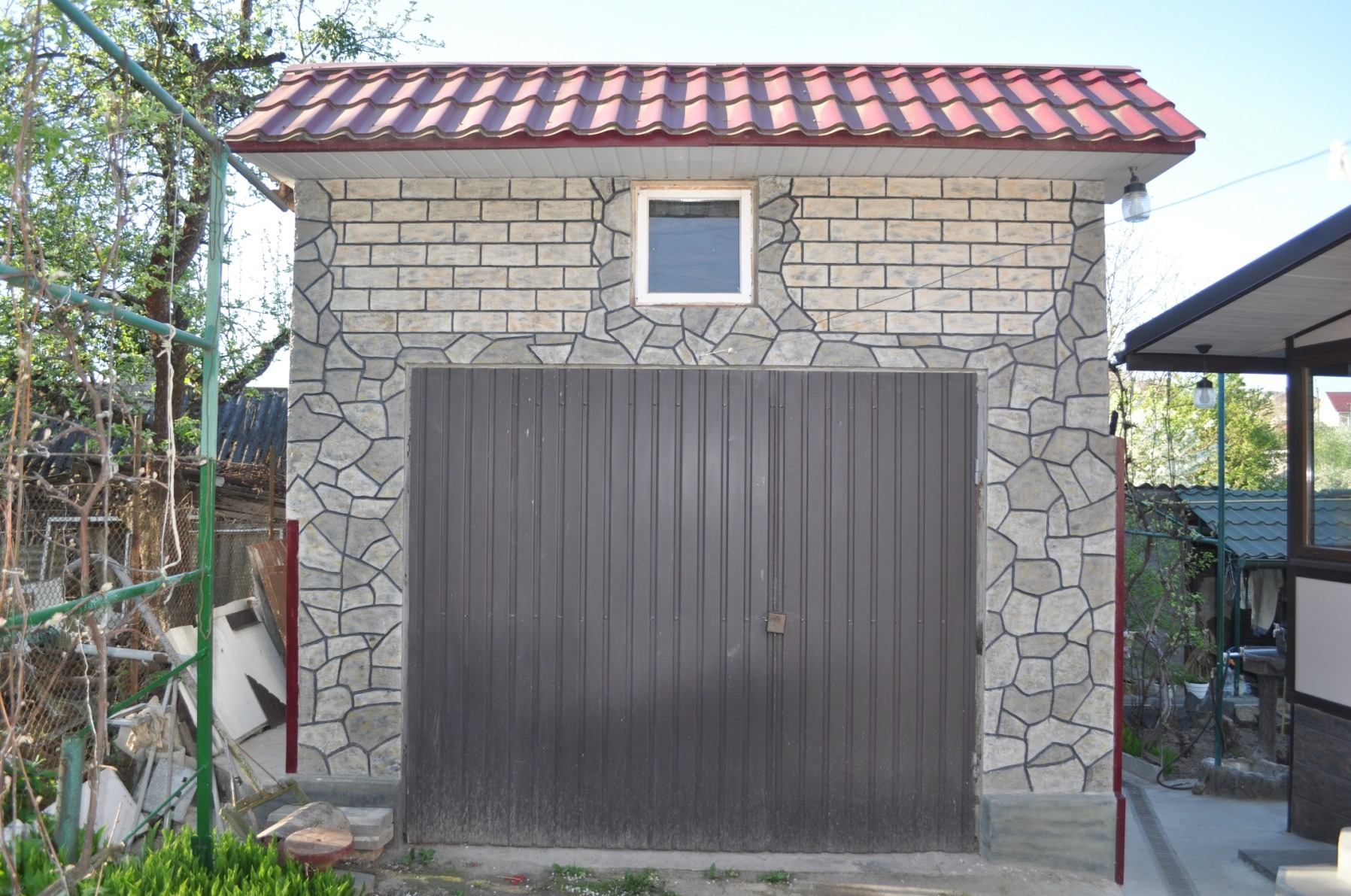 Wykończony garaż z elewacją z kamienia dekoracyjnego i cegły, z brązową bramą segmentową i małym oknem. Czerwona dachówka. Widok z zewnątrz, w tle zieleń.