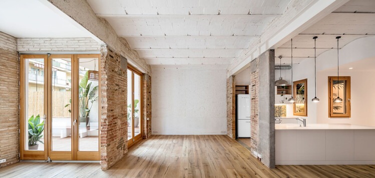 Przestronne wnętrze po renowacji: parkiet, ceglane ściany, białe belki stropowe, okna balkonowe z widokiem na zieleń i otwarta kuchnia z designerskim oświetleniem.