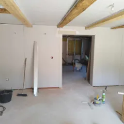 Wnętrze w trakcie prac, ściany obłożone płytami, widoczne drewniane belki stropowe, narzędzia i akcesoria budowlane na podłodze. Otwór drzwiowy bez futryny.