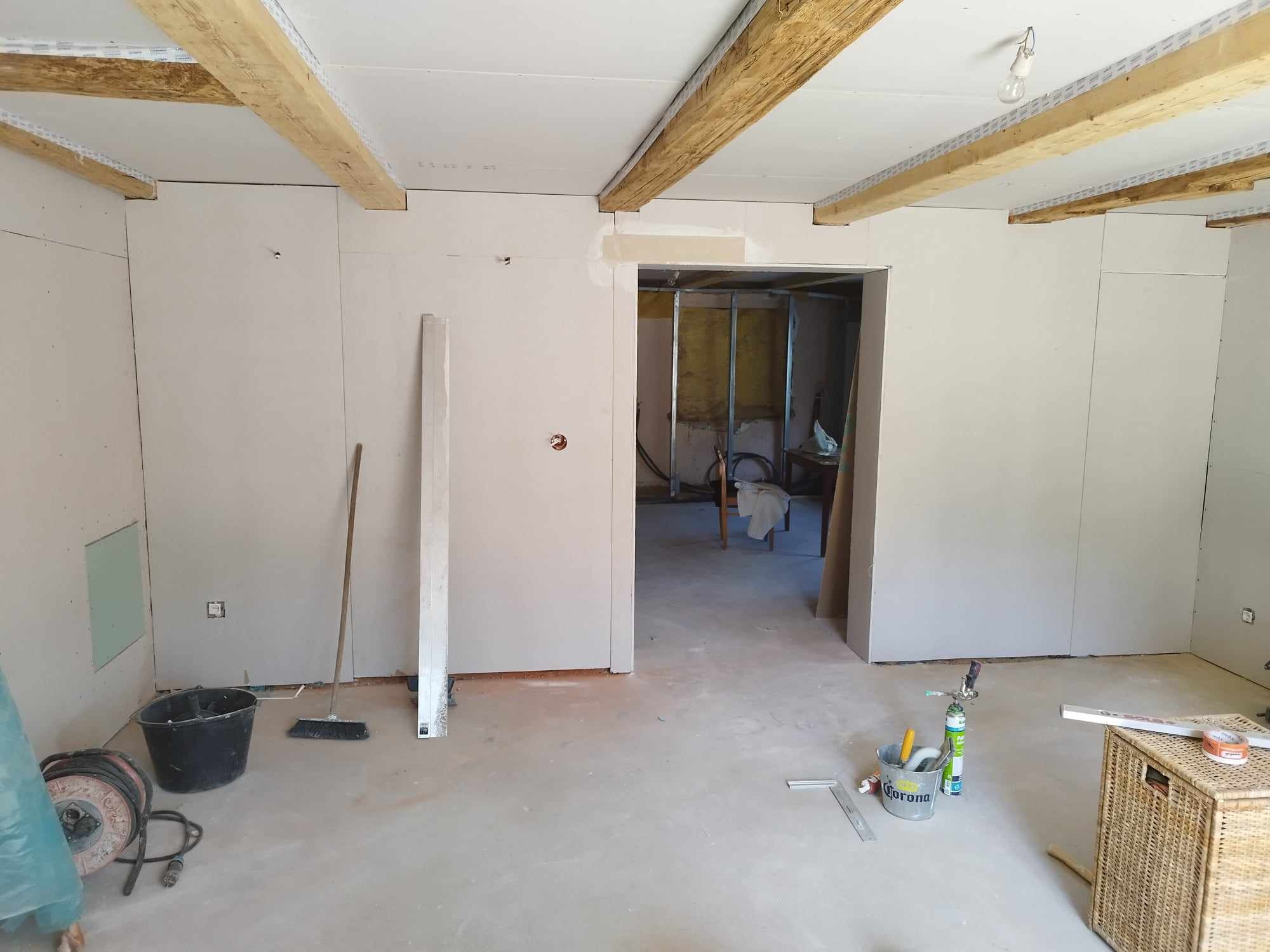 Wnętrze w trakcie prac, ściany obłożone płytami, widoczne drewniane belki stropowe, narzędzia i akcesoria budowlane na podłodze. Otwór drzwiowy bez futryny.