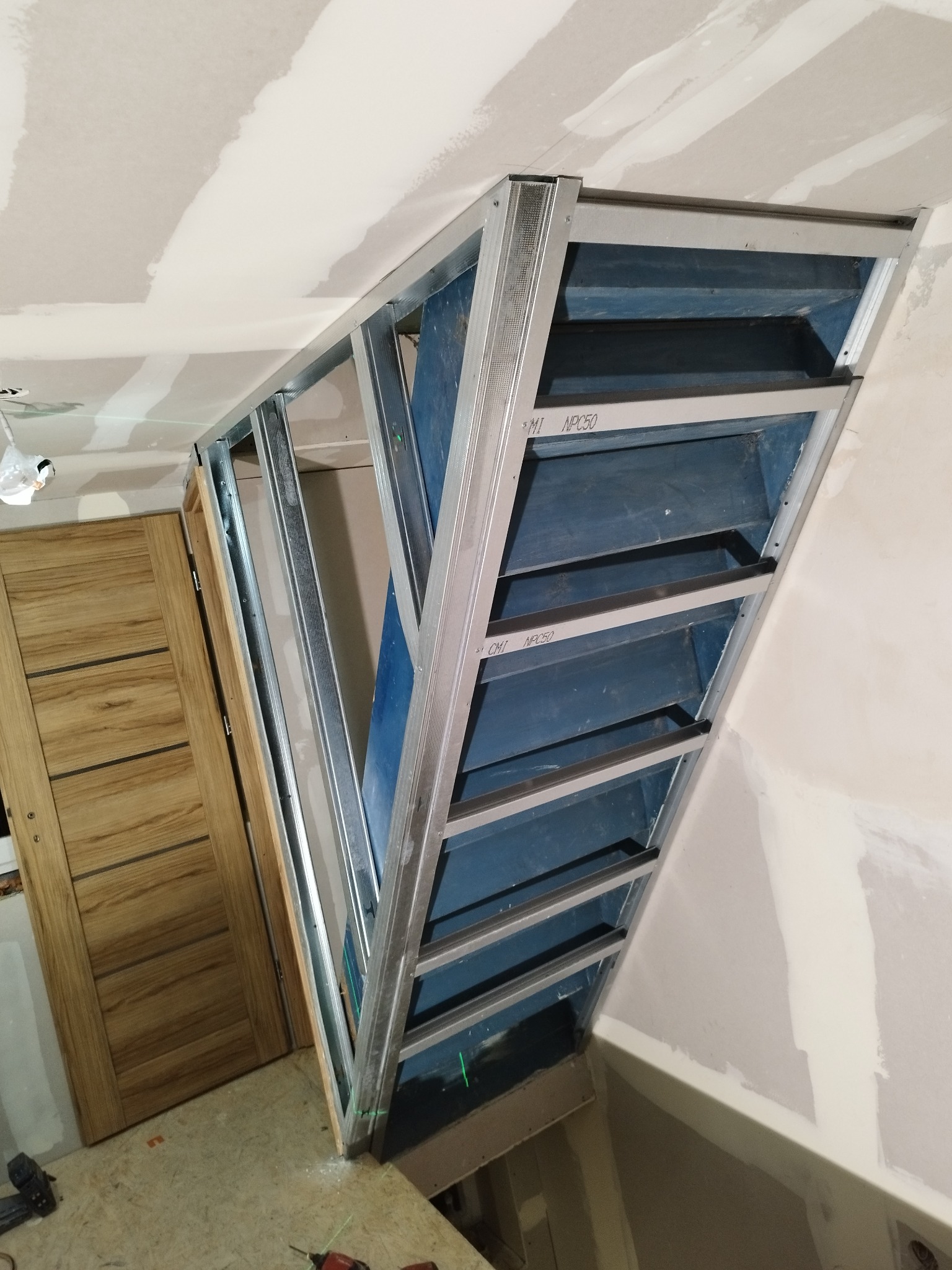 Konstrukcja metalowa pod zabudowę z płyty karton-gips na poddaszu, widoczne profile i niebieskie płyty, drzwi w tle. Etap prac wykończeniowych.