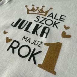 Biała koszulka z okolicznościowym nadrukiem: korona, napis 'ALE SZOK JULKA MAJUŻ ROK' i cyfra '1' z brokatowym efektem, idealna na pierwsze urodziny.