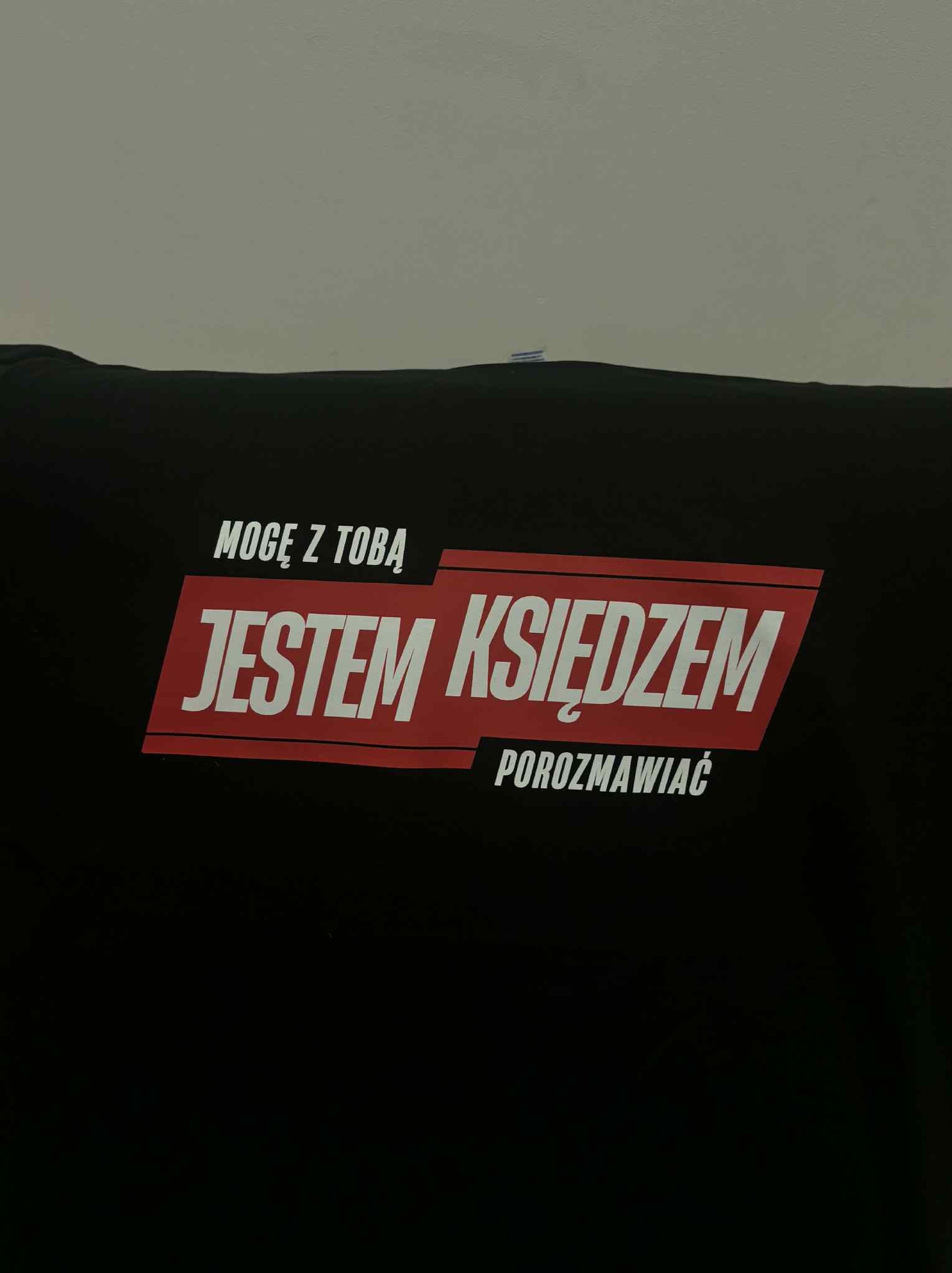 Czarna koszulka z czerwonym nadrukiem: 'Mogę z Tobą JESTEM KSIĘDZEM Porozmawiać'. Nietypowy humorystyczny napis na odzieży.