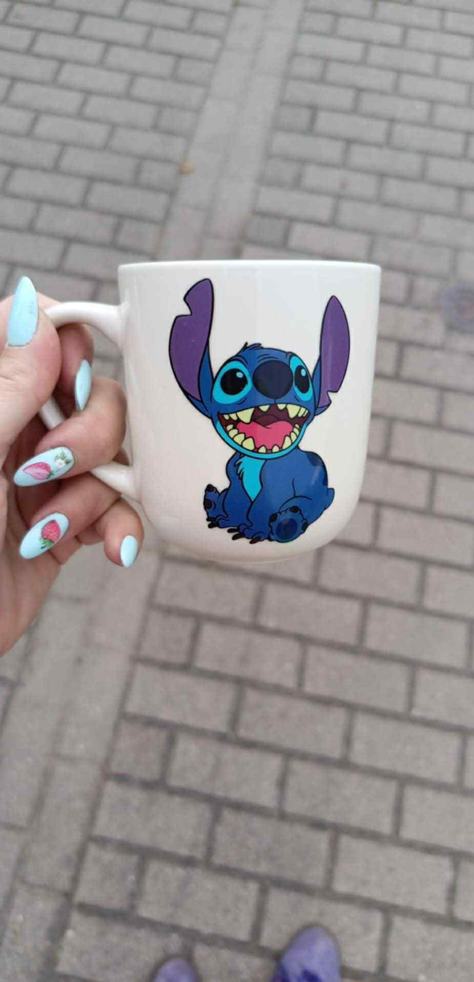 Biały kubek z nadrukiem Stitcha trzymany w dłoni z błękitnymi paznokciami zdobionymi motywem truskawek na tle szarej kostki brukowej.