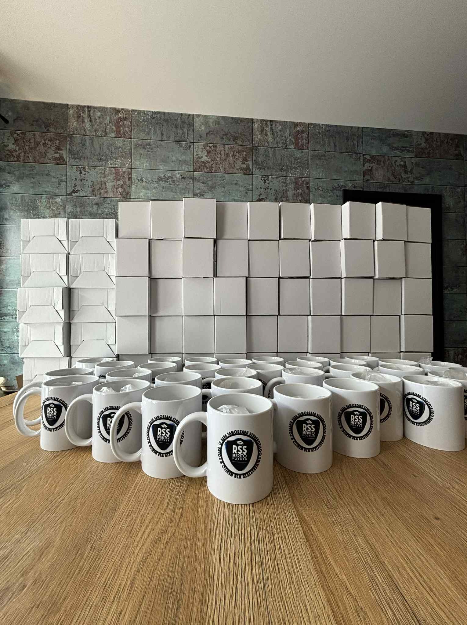 Rząd białych kubków z czarnym nadrukiem logo 'RSS Merito Poznań' na drewnianym stole, za nimi stosy białych pudełek, tworzące kompozycję.