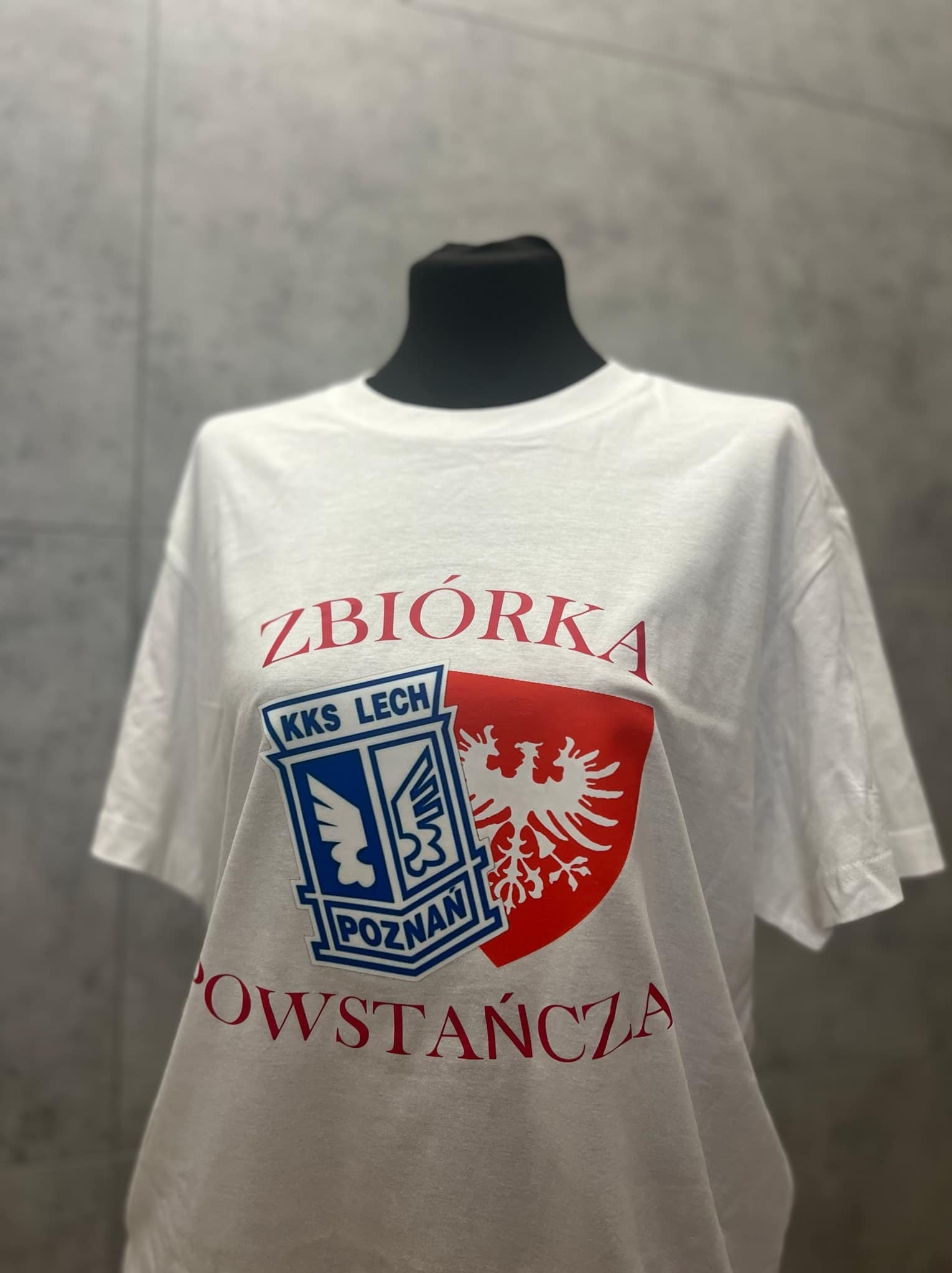 Biała koszulka z czerwono-niebieskim nadrukiem: herb KKS Lech Poznań, orzeł i napis 'Zbiórka Powstańcza', prezentowana na manekinie na tle szarej ściany.