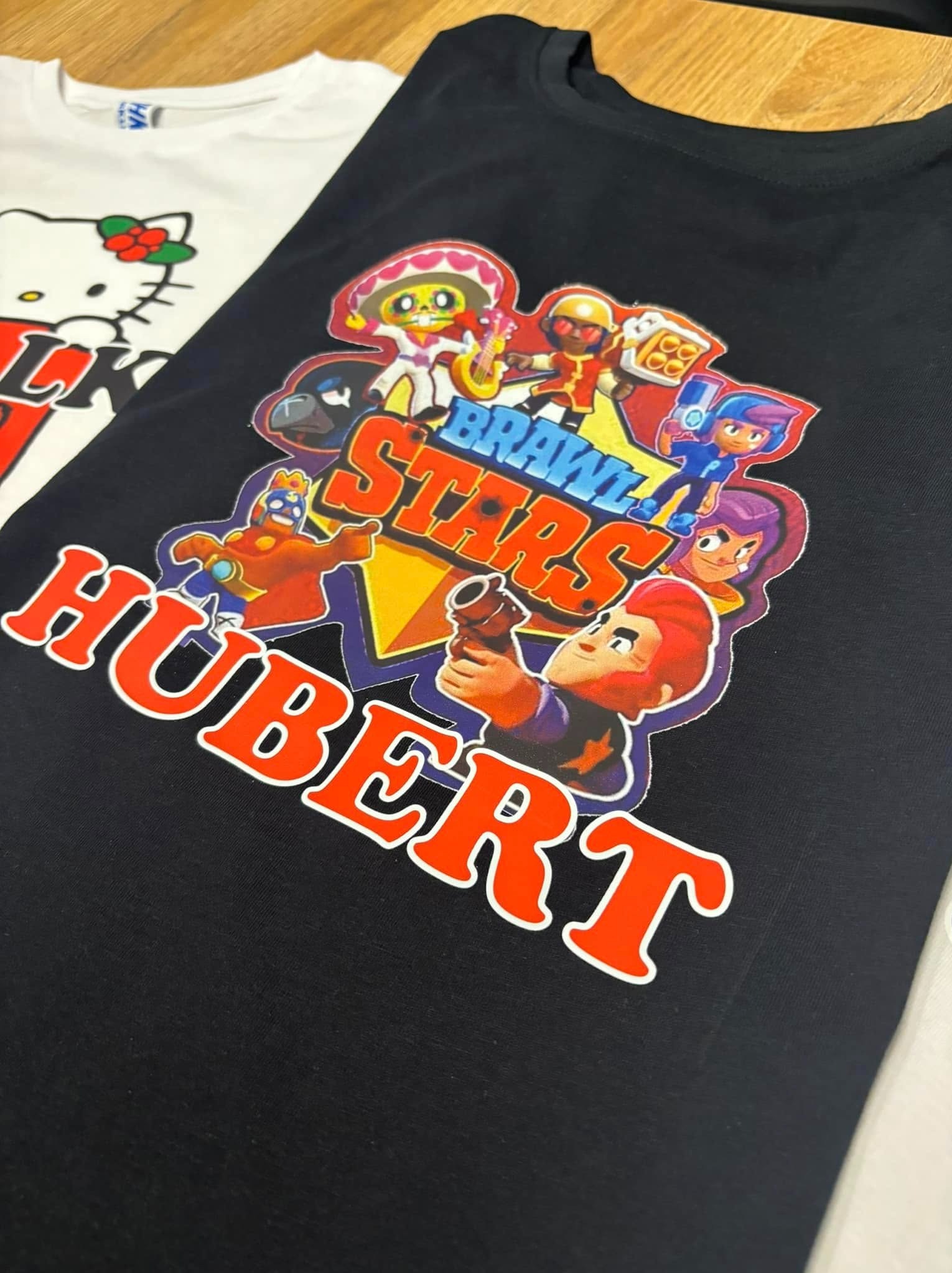 Czarna koszulka z nadrukiem postaci z gry Brawl Stars i imieniem Hubert. Obok biała koszulka z Hello Kitty. Ujęcie z bliska, na drewnianym blacie.