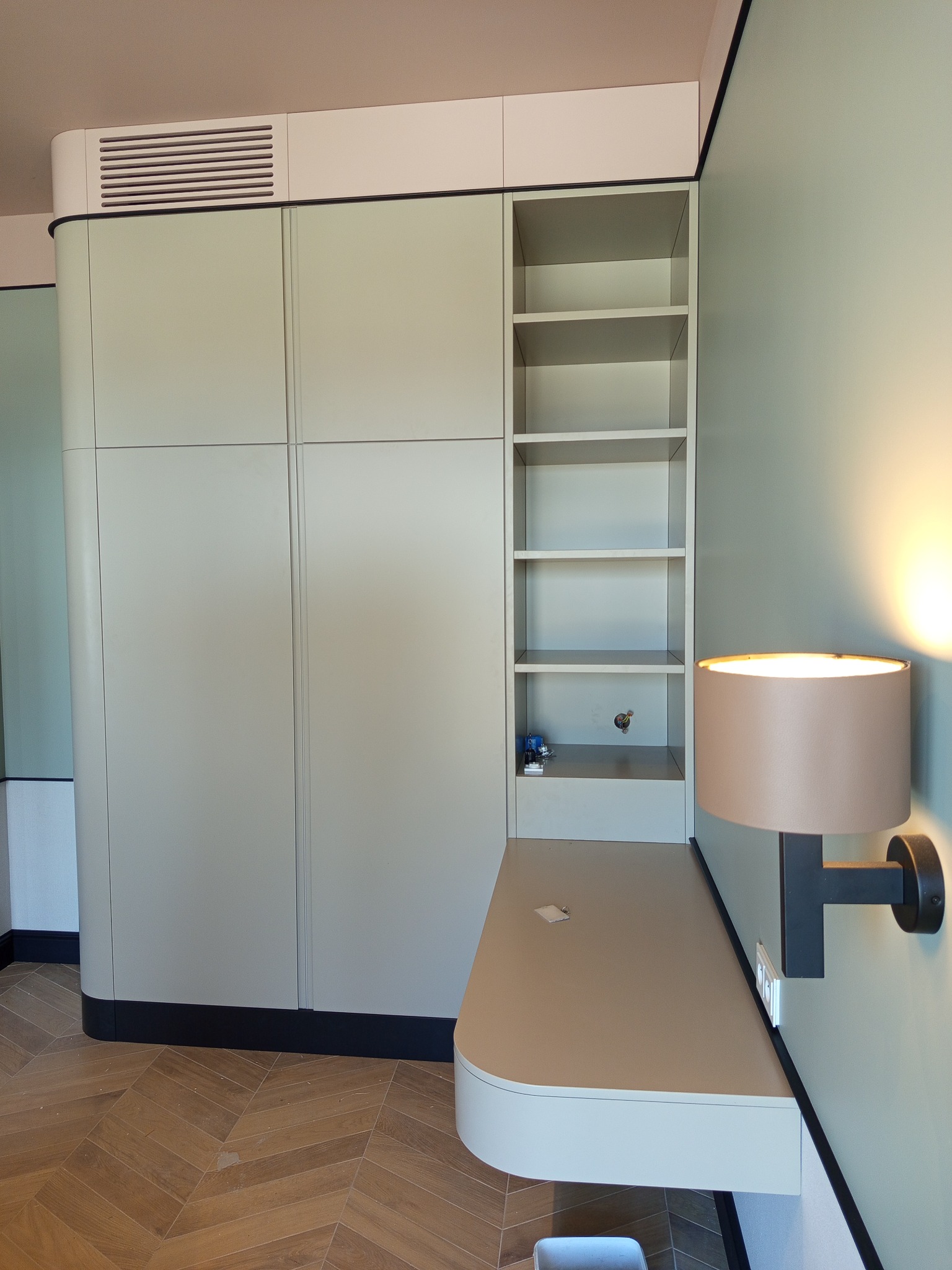 Jasnozielona zabudowa meblowa z szafą, półkami i blatem, oświetlona lampą ścienną. Widoczne zaokrąglone krawędzie i parkiet w jodełkę.