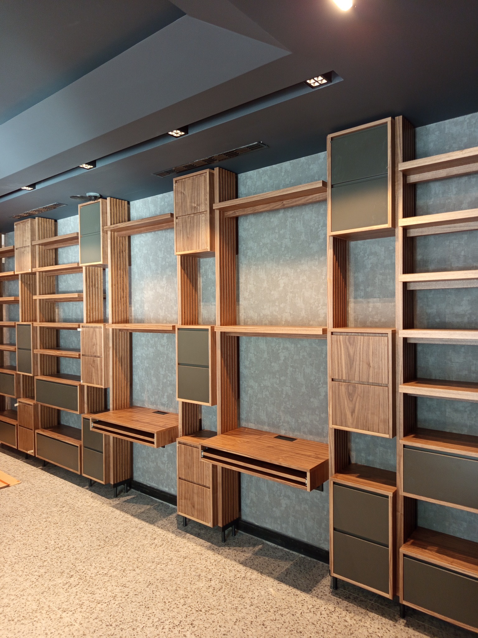 Nowoczesny system regałów i szafek z drewnianymi elementami i szarymi frontami, zamontowany na ścianie z szarą tapetą. Widoczne biurka i oświetlenie sufitowe.