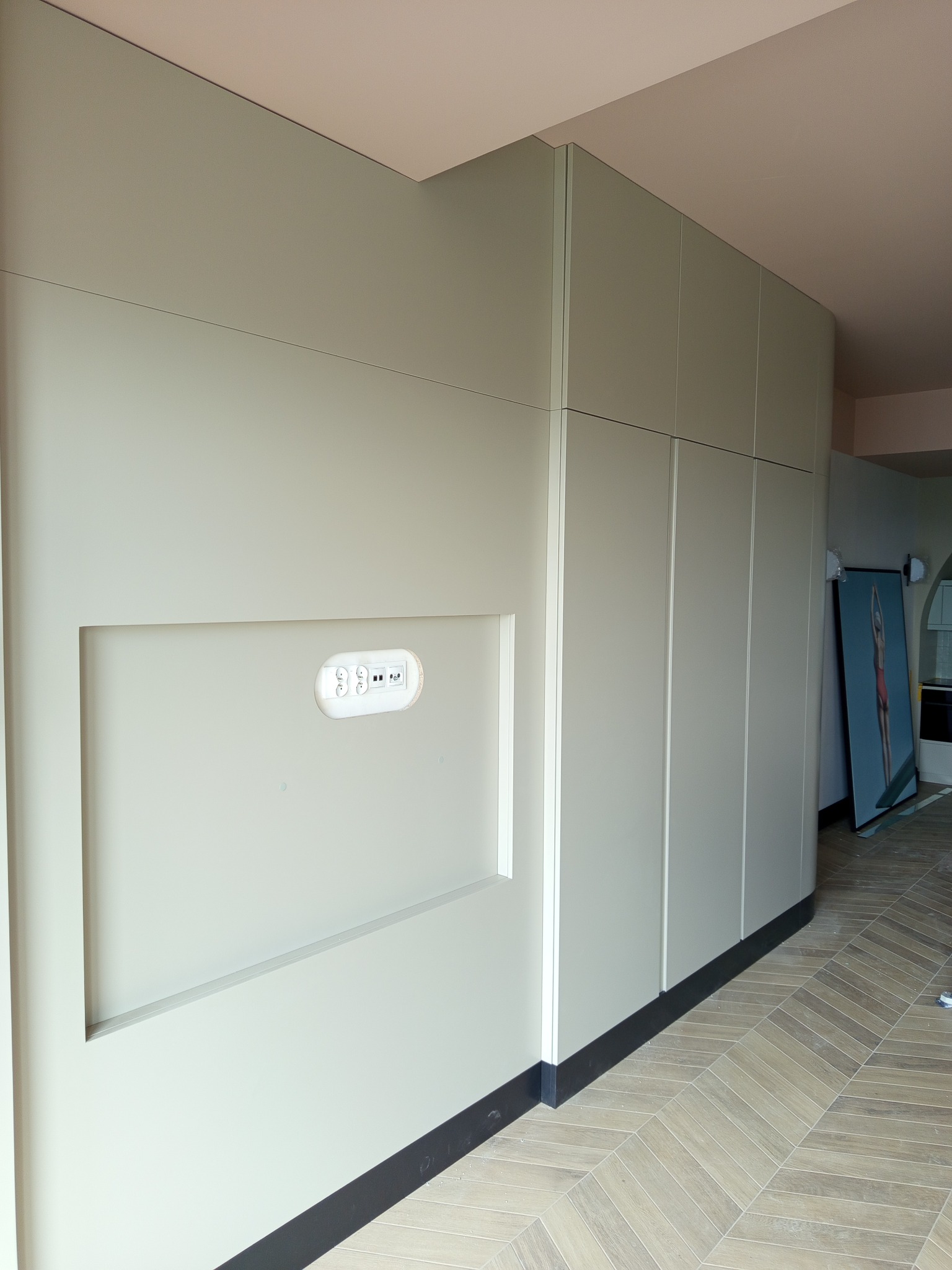 Nowoczesna zabudowa ścienna w odcieniach szarości z wnęką na multimedia i gniazdkami, uzupełniona o obraz oparty o ścianę na parkiecie w jodełkę.