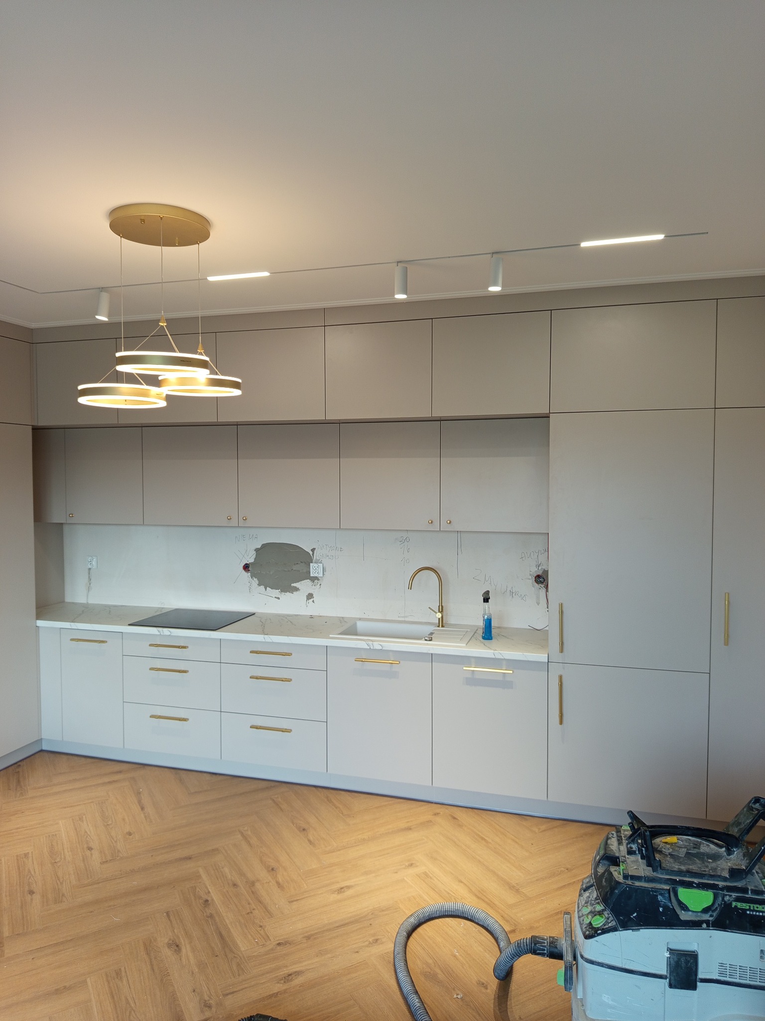 Jasna kuchnia z złotymi uchwytami i lampami, widoczne ślady po montażu na ścianie, odkurzacz na podłodze. Parkiet w jodełkę.
