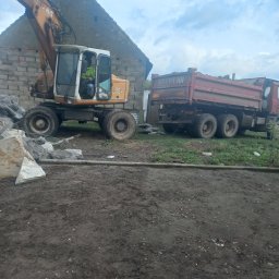 Sprzedaż żwiru I piasku transport od 1t do 20t - Żółta koparka kołowa obok czerwonej ciężarówki, prawdopodobnie podczas prac rozbiórkowych lub porządkowych na terenie budowy, z gruzem w tle.