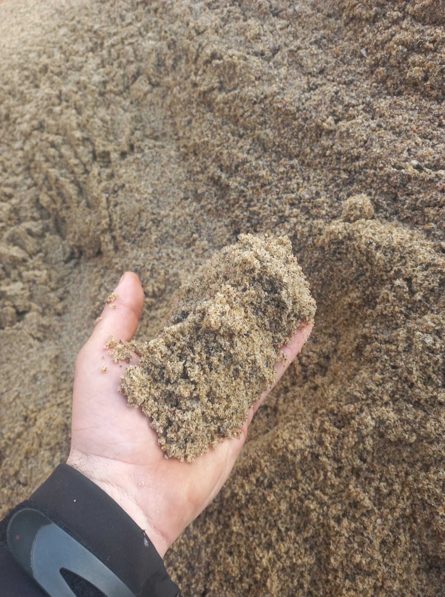 Dłoń w czarnej rękawicy trzyma piasek na tle dużej pryzmy piasku o nierównej fakturze i naturalnym oświetleniu.