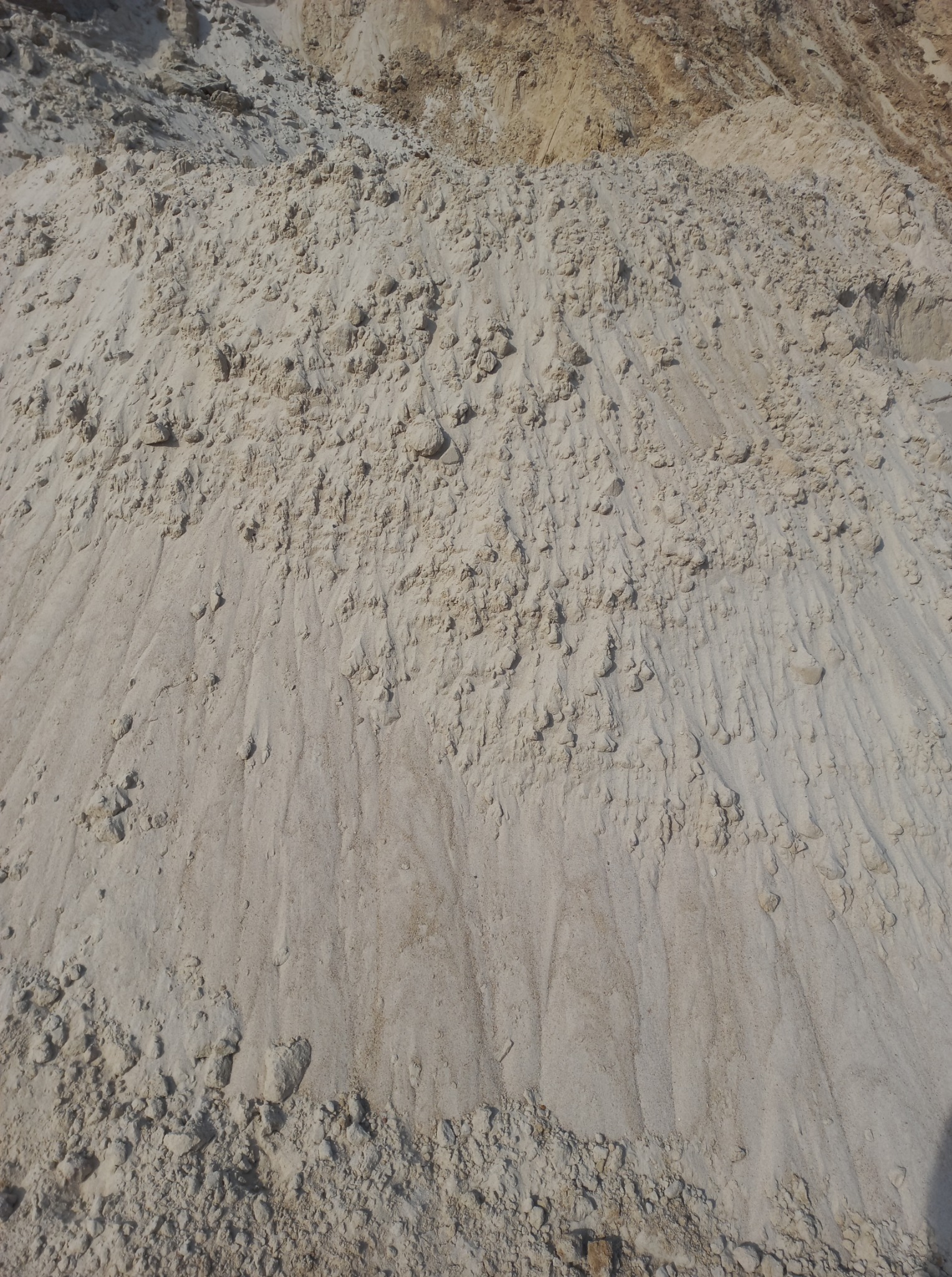 Jasny piasek z niewielkimi kamieniami, ułożony w pryzmę. Widoczne ślady po zsypywaniu i nierówna powierzchnia. U góry ciemniejsza warstwa ziemi.