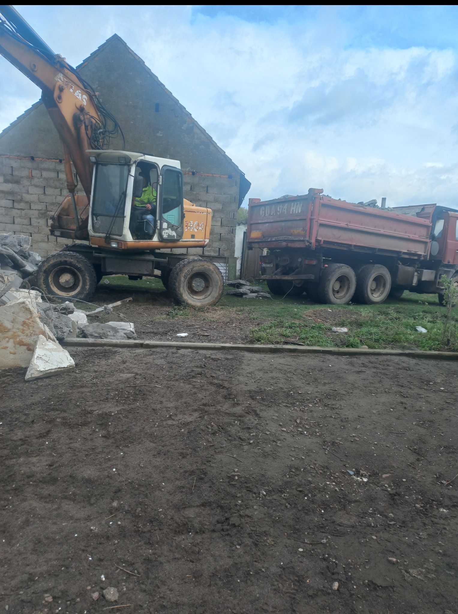 Żółta koparka kołowa obok czerwonej ciężarówki, prawdopodobnie podczas prac rozbiórkowych lub porządkowych na terenie budowy, z gruzem w tle.