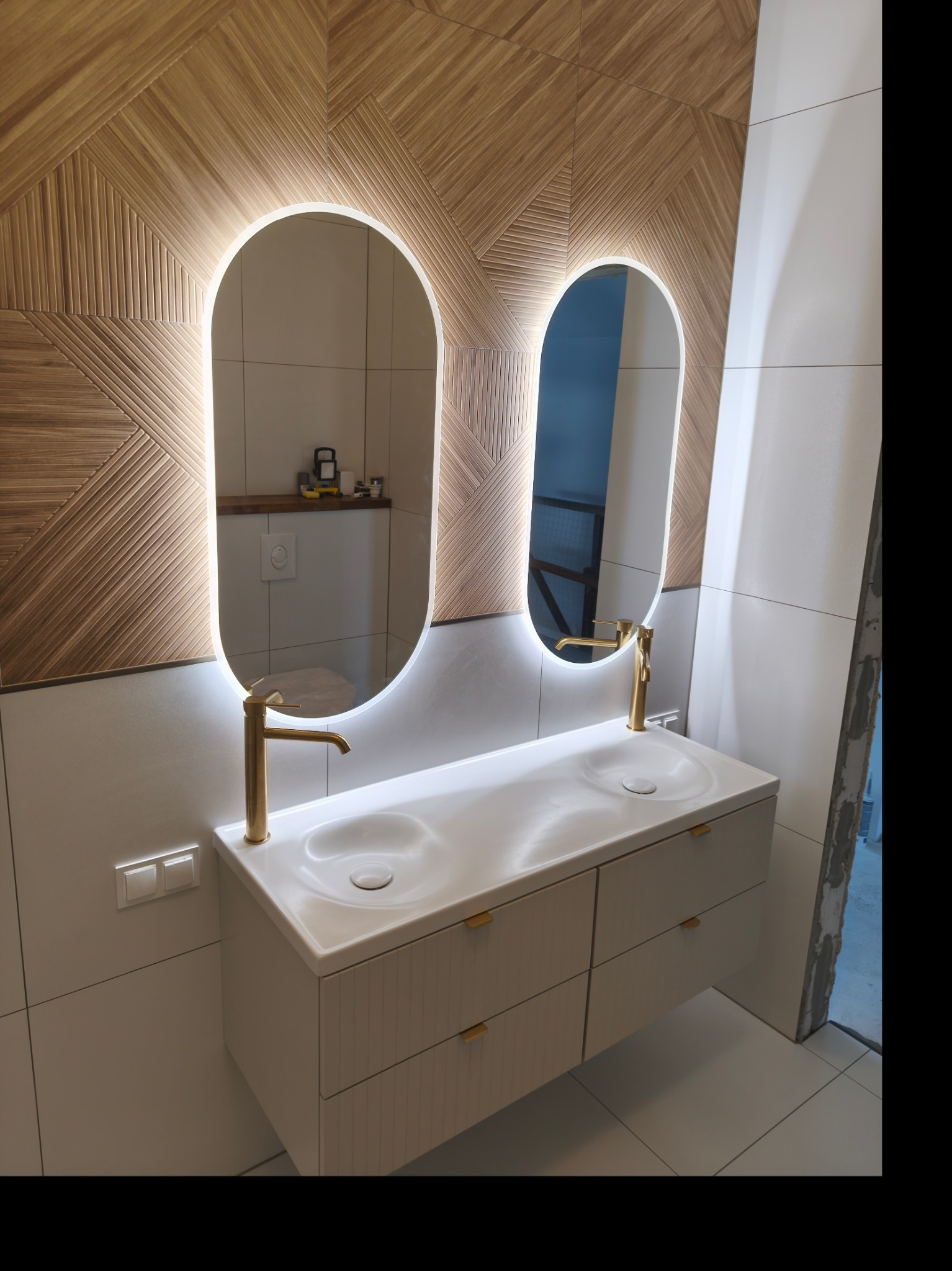 Nowoczesna łazienka z podwójną umywalką, złotą armaturą i dekoracyjną ścianą z drewnopodobnych płytek ułożonych we wzór. Owalne lustra z podświetleniem LED.