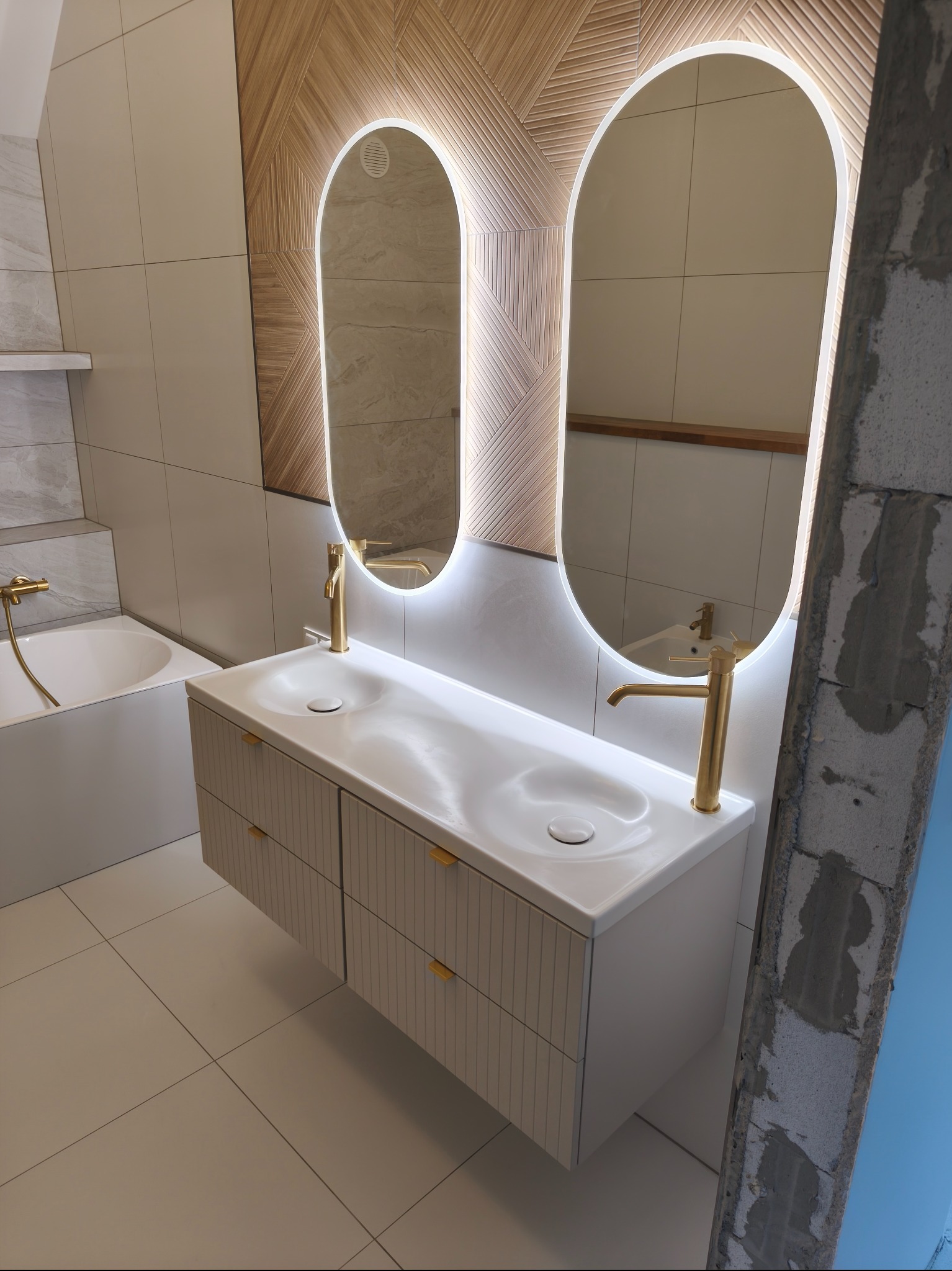 Nowoczesna łazienka z owalnymi lustrami podświetlanymi LED, białą szafką z umywalką, złotą armaturą i drewnianą mozaiką na ścianie. Częściowo widoczny surowy mur.