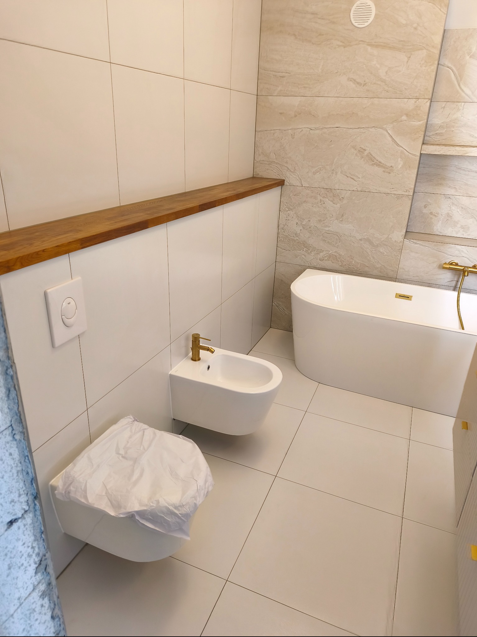 Nowoczesna łazienka z wanną, bidetem i toaletą w białej kolorystyce, wykończona płytkami w odcieniach beżu i drewna. Złota armatura dodaje elegancji.