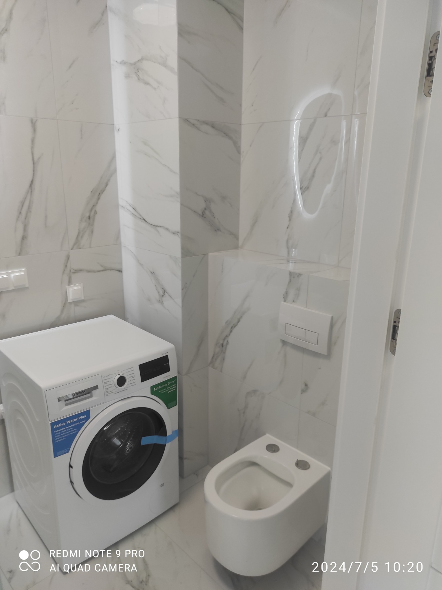 Wnętrze łazienki po remoncie: białe marmurowe płytki, podwieszana toaleta i pralka Bosch. Widoczny fragment białych drzwi i przycisk spłuczki.