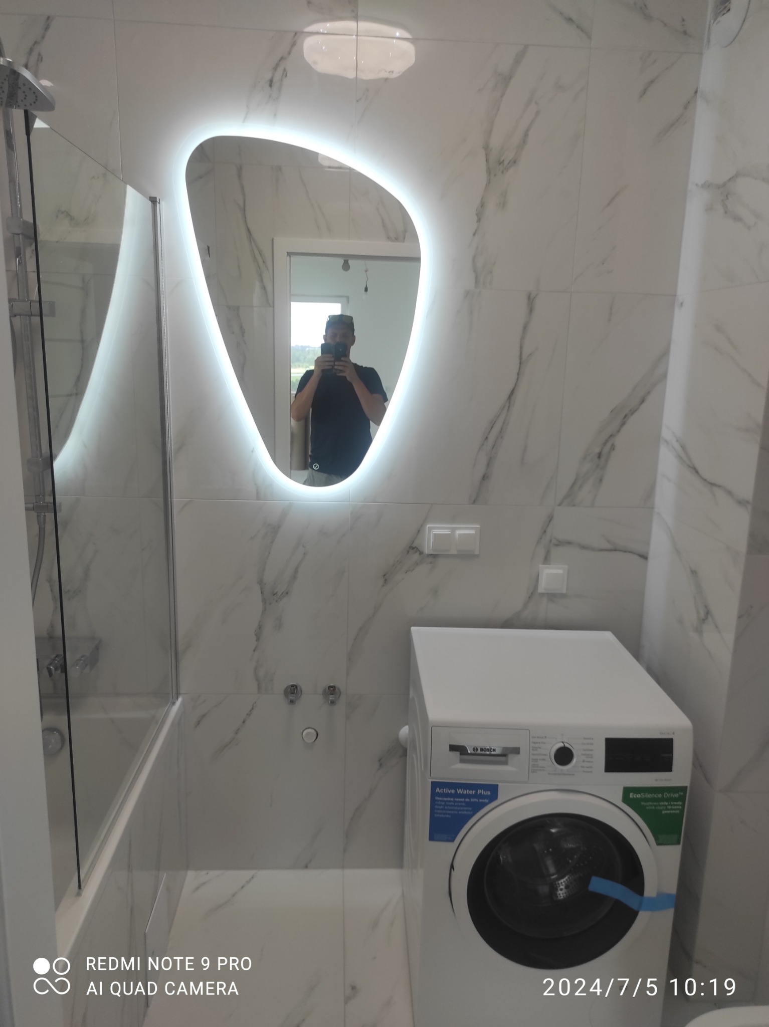Wykończona łazienka w bieli: marmurowe płytki, designerskie lustro z podświetleniem, pralka Bosch, kabina prysznicowa. Widoczne detale wykończenia i odbicie osoby w lustrze.