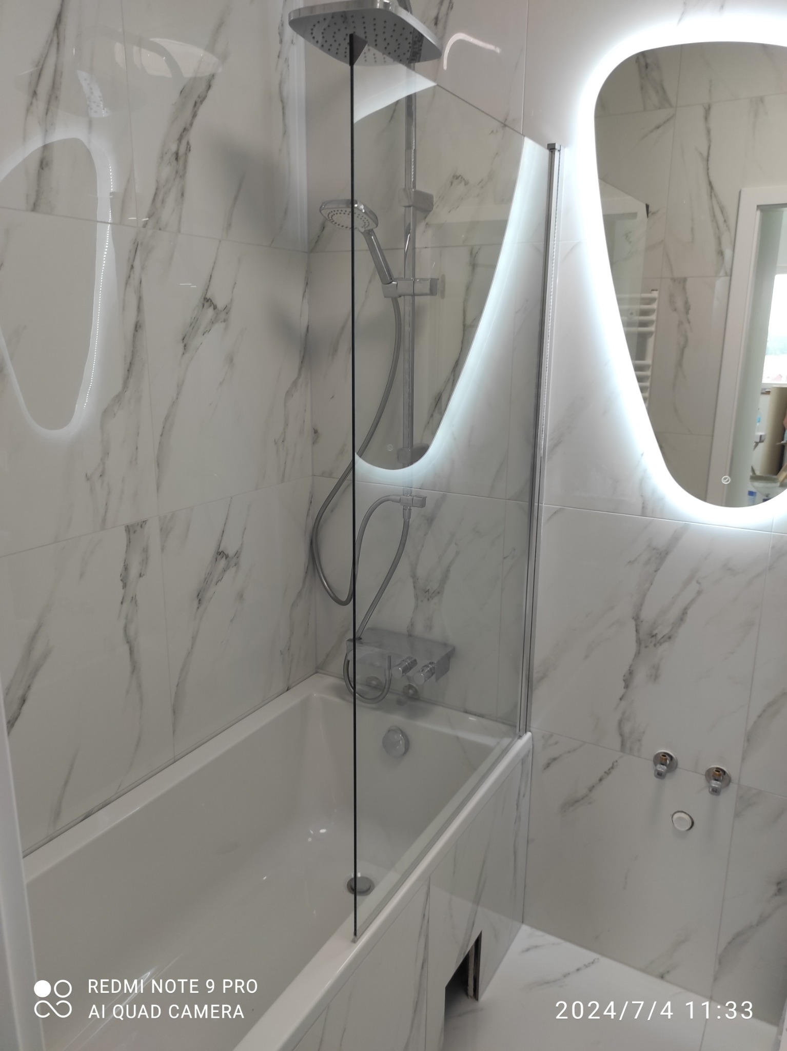 Biała łazienka z wanną i kabiną prysznicową, wykończona płytkami imitującymi marmur. Podświetlane lustro nadaje wnętrzu nowoczesny charakter.