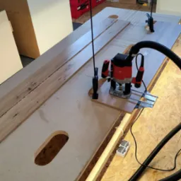 Frezarka w trakcie pracy nad obróbką drewna na stole warsztatowym, widoczne elementy mocujące i system odsysania pyłu. W tle skrzynki narzędziowe i regał.