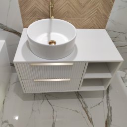 almost heaven Piotr Antoni Witkowski - Biała szafka łazienkowa z umywalką nablatową i złotą armaturą. Fronty szuflad z ryflowanym wzorem, marmurowe płytki na podłodze i ścianie.