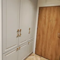 Elegancka, szara szafa wnękowa z złotymi uchwytami w kształcie muszli, na tle drewnianej podłogi w jodełkę i drzwi. Minimalistyczny design.