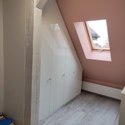 almost heaven Piotr Antoni Witkowski - Biała szafa wnękowa na poddaszu, z drewnianą belką i oknem dachowym. Widoczna podłoga z paneli i fragment grzejnika. U dołu narzędzia i farby.