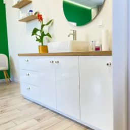 Biała szafka łazienkowa z blatem z drewna, okrągłe lustro, kwiat w doniczce i kosmetyki. Całość w nowoczesnym stylu.