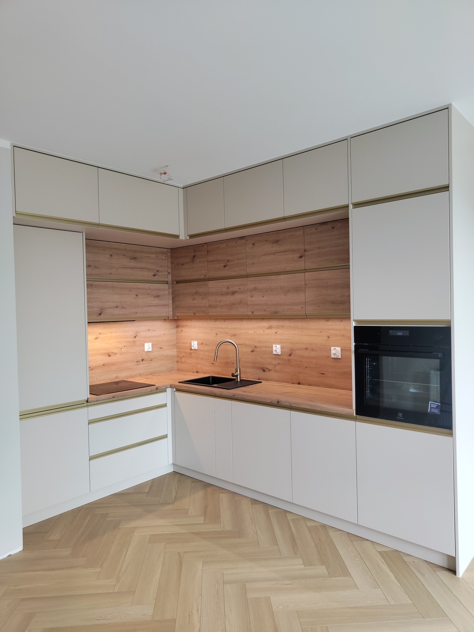 Nowoczesna kuchnia z białymi szafkami, drewnianym blatem i złotymi akcentami. Na podłodze parkiet ułożony w jodełkę. Widoczny piekarnik i płyta indukcyjna.