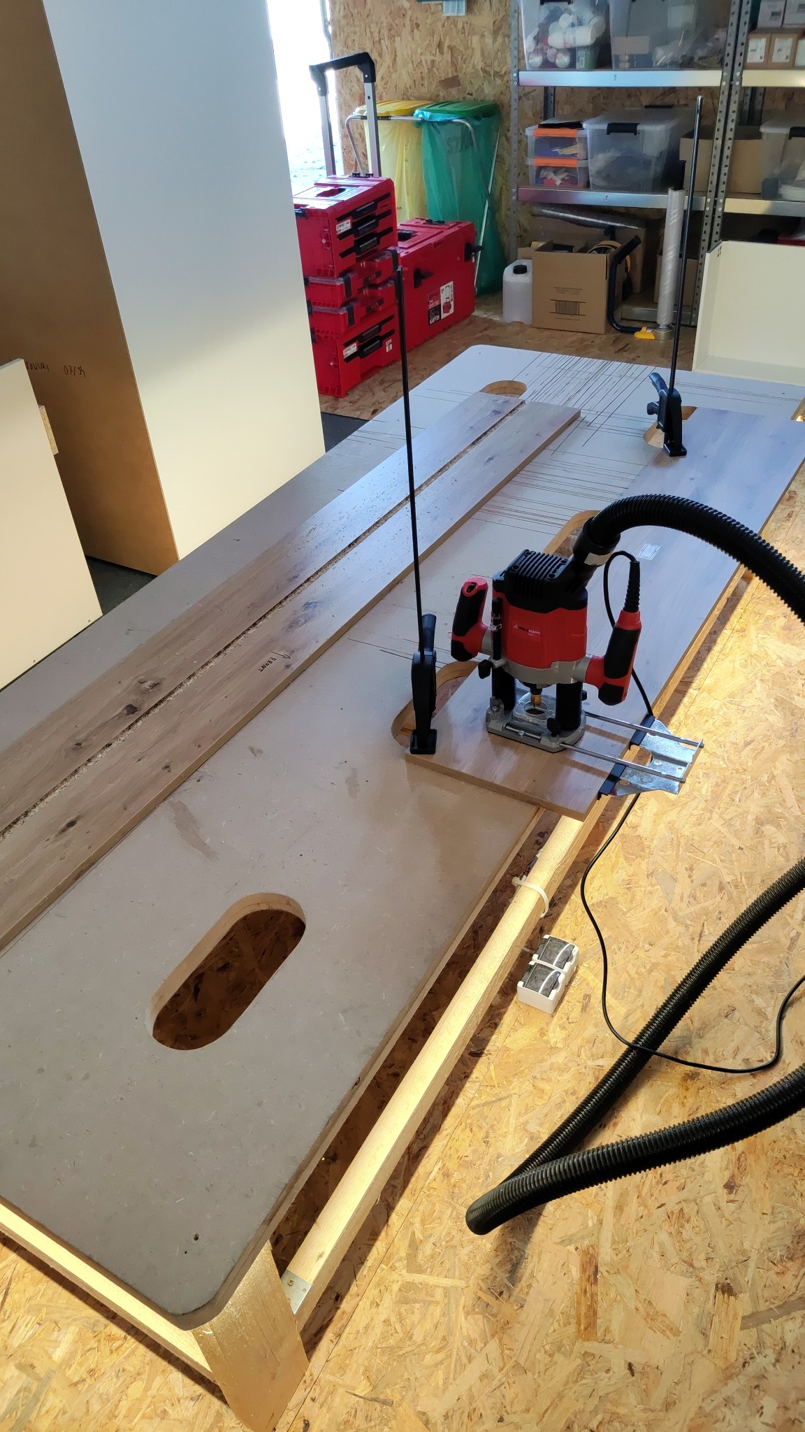 Frezarka w trakcie pracy nad obróbką drewna na stole warsztatowym, widoczne elementy mocujące i system odsysania pyłu. W tle skrzynki narzędziowe i regał.