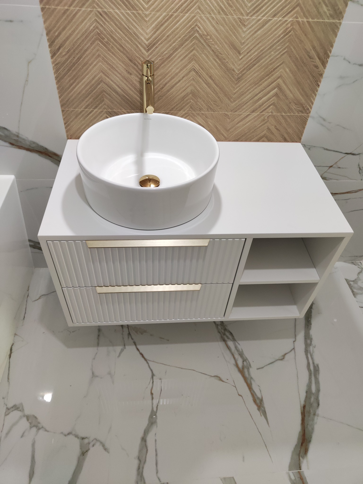 Biała szafka łazienkowa z umywalką nablatową i złotą armaturą. Fronty szuflad z ryflowanym wzorem, marmurowe płytki na podłodze i ścianie.