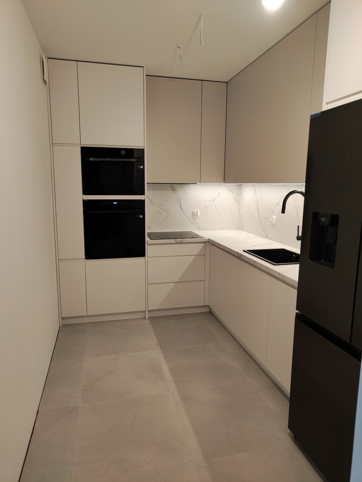 Nowoczesna kuchnia z białymi frontami, czarnym piekarnikiem i lodówką. Marmurowy blat i oświetlenie LED dodają elegancji. Minimalistyczny design.