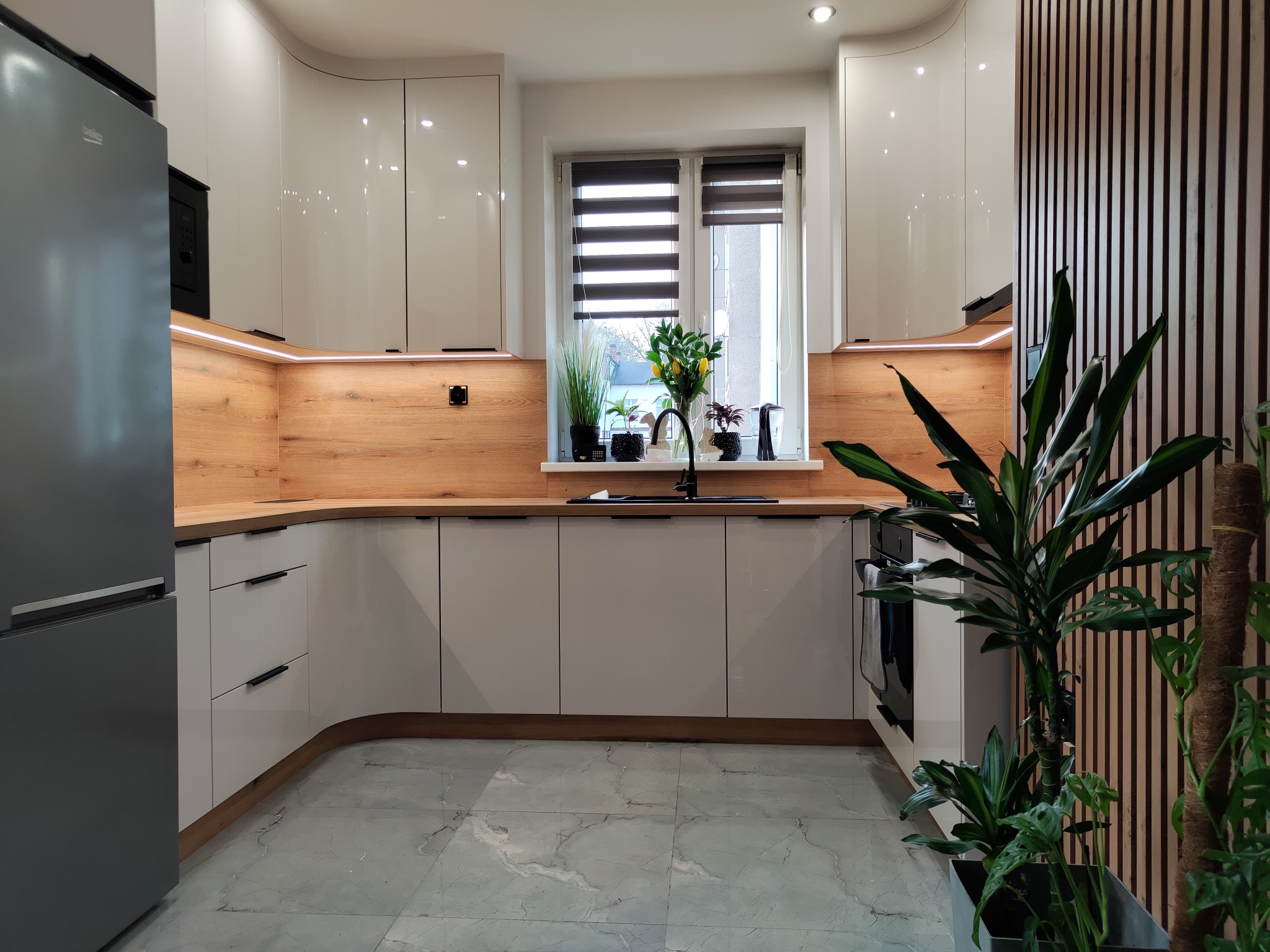 Jasna kuchnia z białymi, zaokrąglonymi szafkami, drewnianym blatem i panelami ściennymi. Rośliny w oknie i na podłodze dodają naturalnego akcentu.