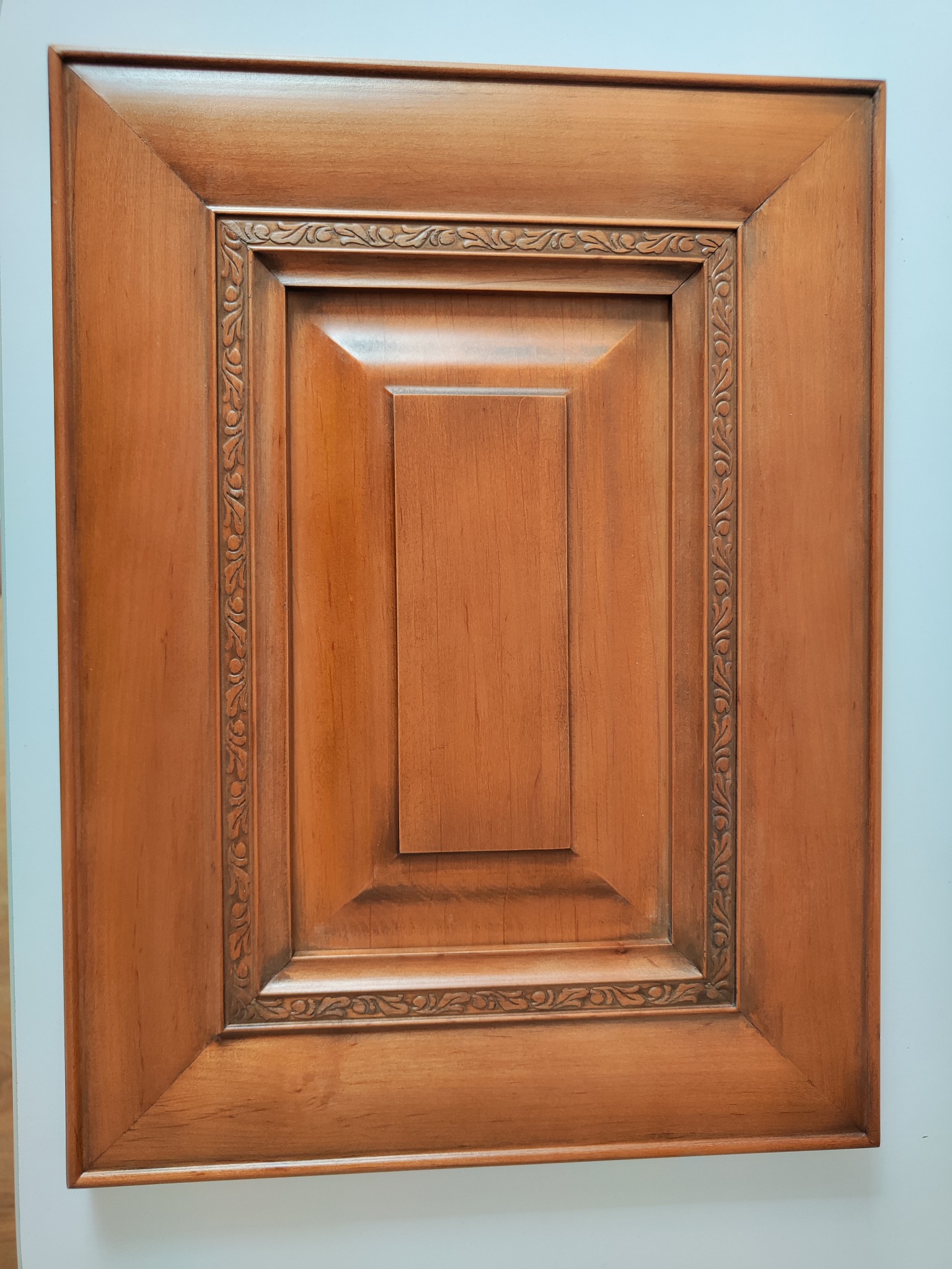 Drewniane drzwiczki szafki z dekoracyjnym frezem, w ciepłym odcieniu brązu, ukazane z bliska. Widoczna struktura drewna i detale wykończenia.