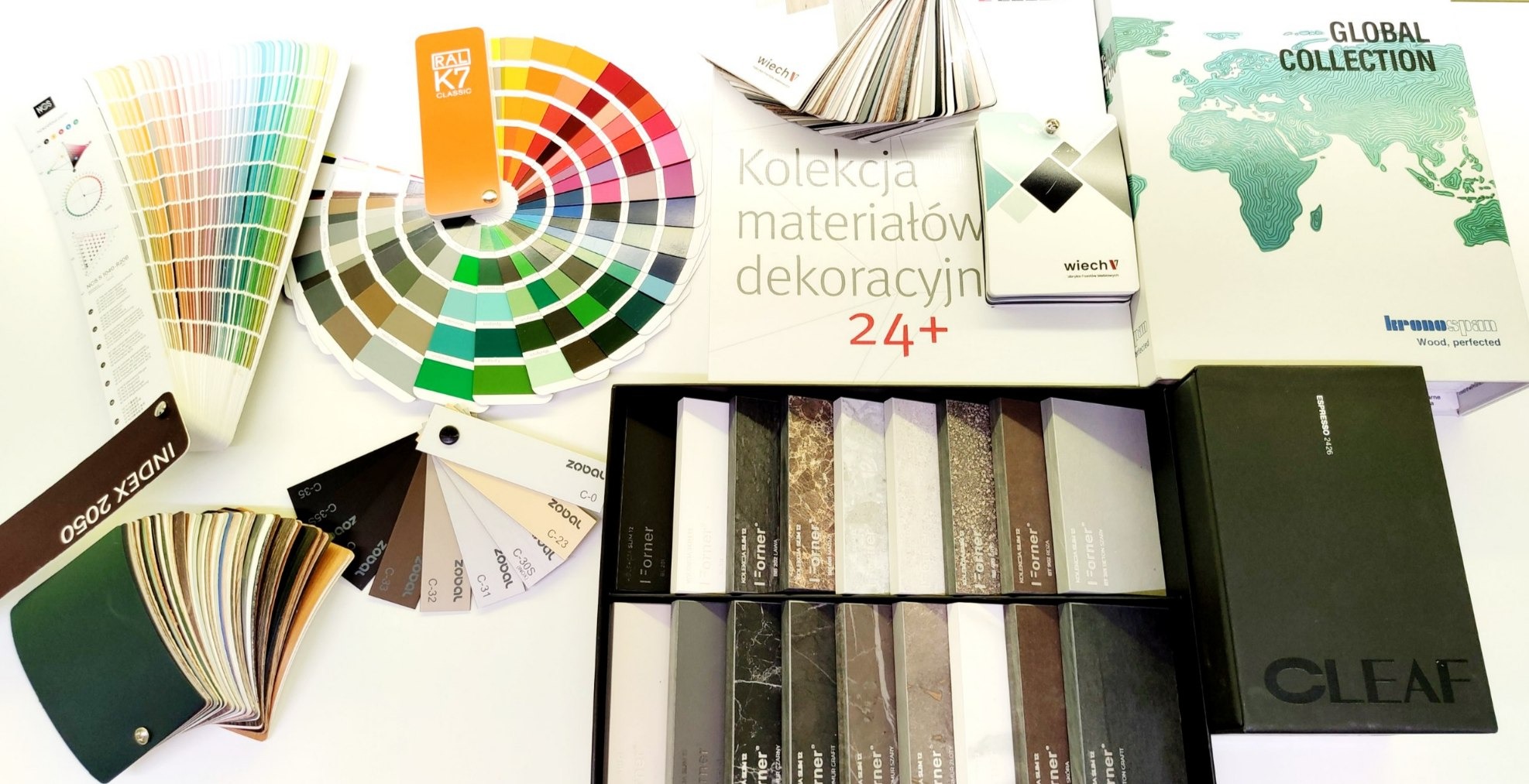 Palety barw RAL, próbniki fornirów i materiałów dekoracyjnych, katalogi Kronospan i Cleaf na białym stole. Prezentacja różnorodności kolorów i tekstur.