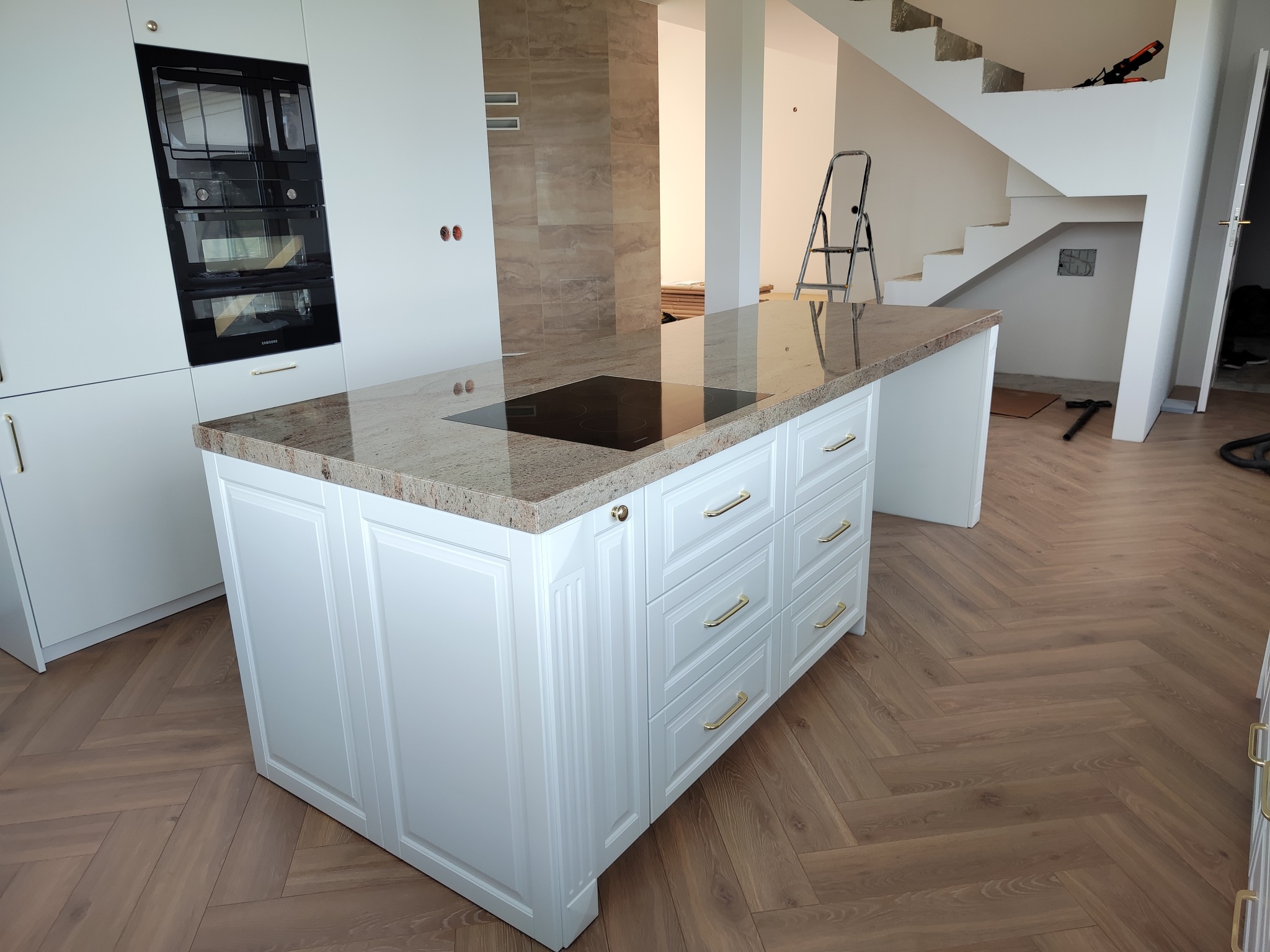 Biała wyspa kuchenna z kamiennym blatem i złotymi uchwytami, w tle zabudowa kuchenna i schody w surowym stanie, parkiet w jodełkę.