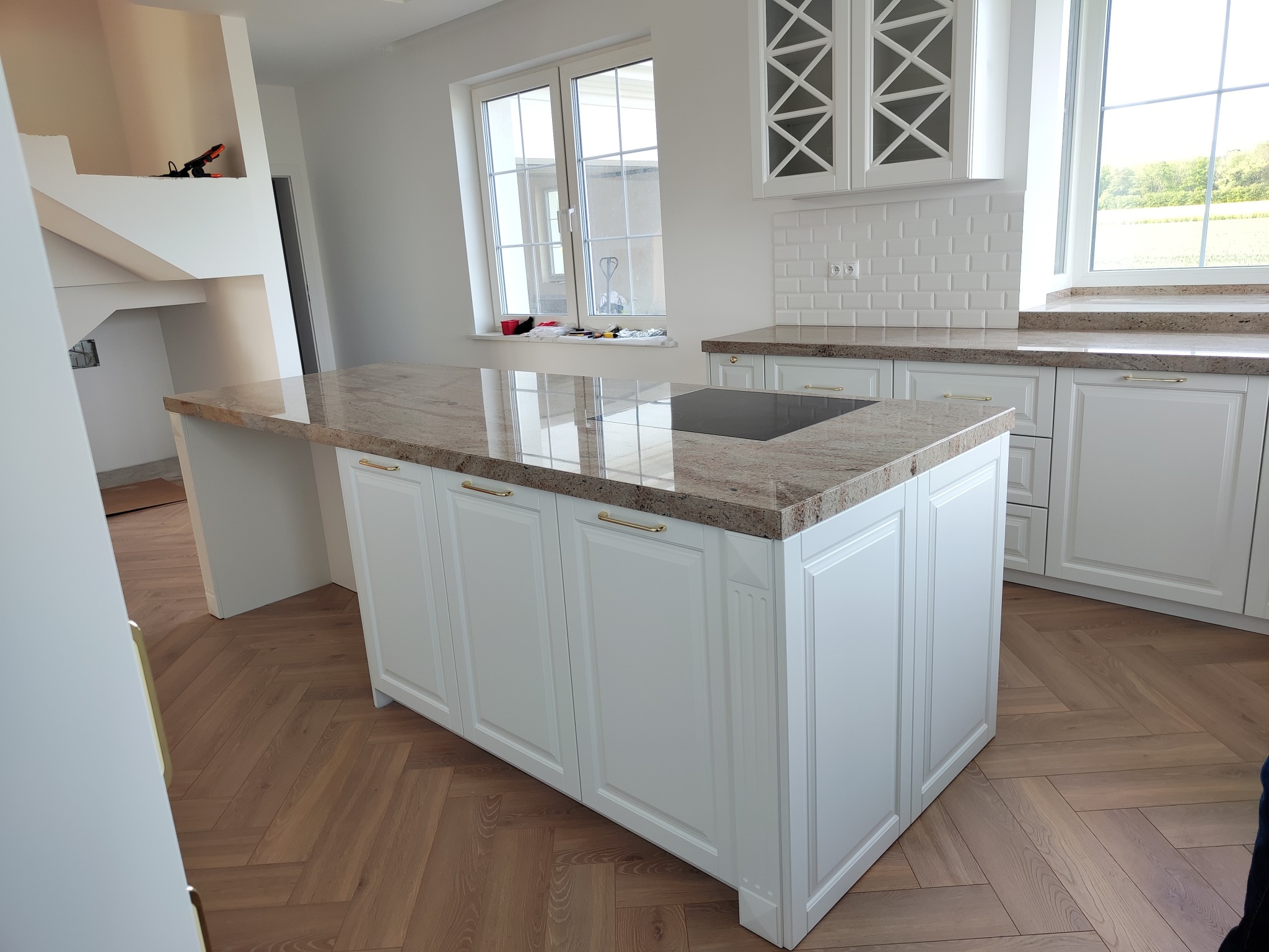 Jasna kuchnia z wyspą i blatem z kamienia. Białe szafki z frezowanymi frontami. Parkiet ułożony w jodełkę. Okno z widokiem na zieleń. Nowoczesny design.