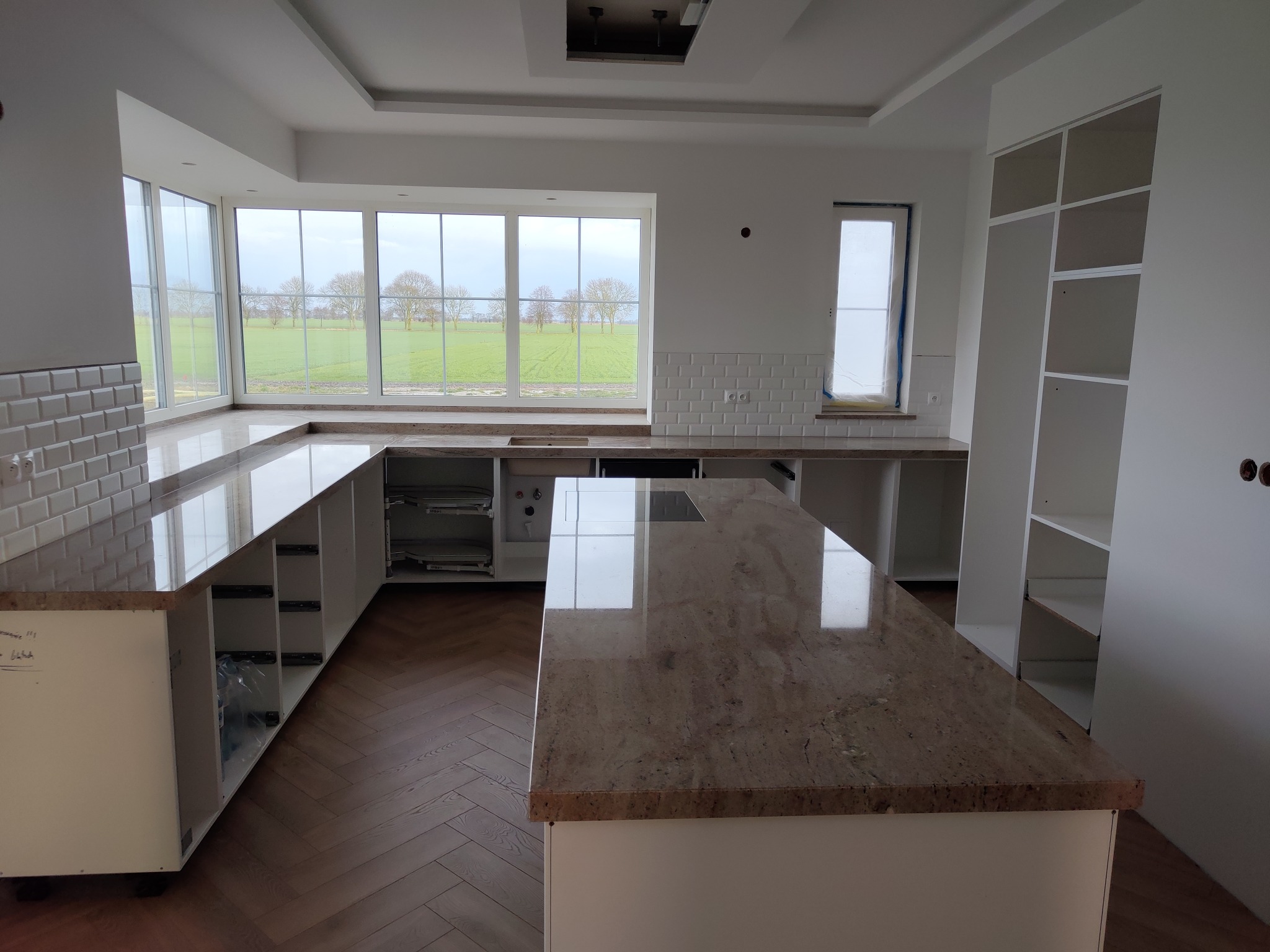 Jasna, nowoczesna kuchnia w trakcie montażu: wyspa z kamiennym blatem, okno z widokiem na pola, białe płytki i podłoga w jodełkę. Widoczne niedokończone szafki.