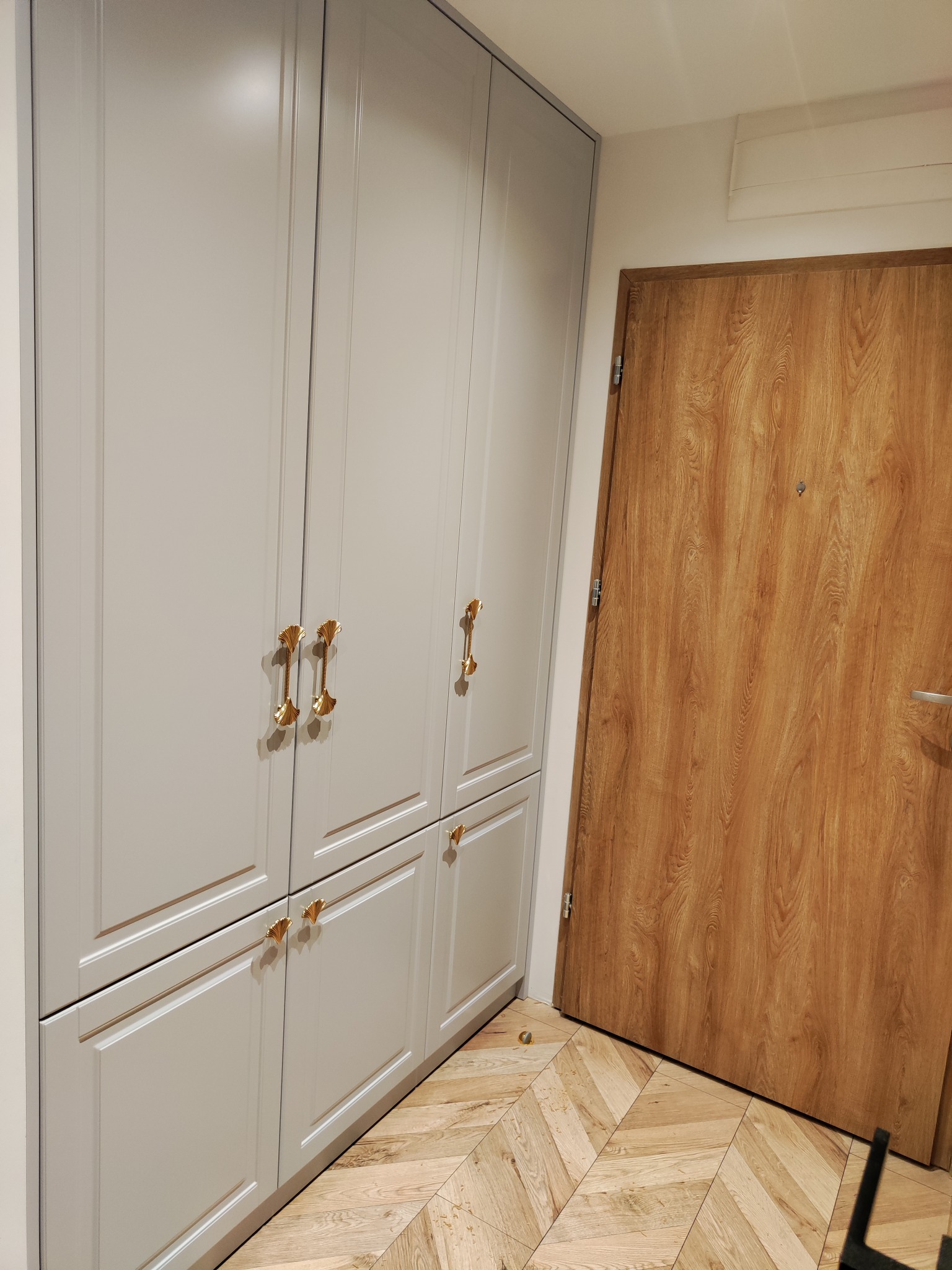 Elegancka, szara szafa wnękowa z złotymi uchwytami w kształcie muszli, na tle drewnianej podłogi w jodełkę i drzwi. Minimalistyczny design.