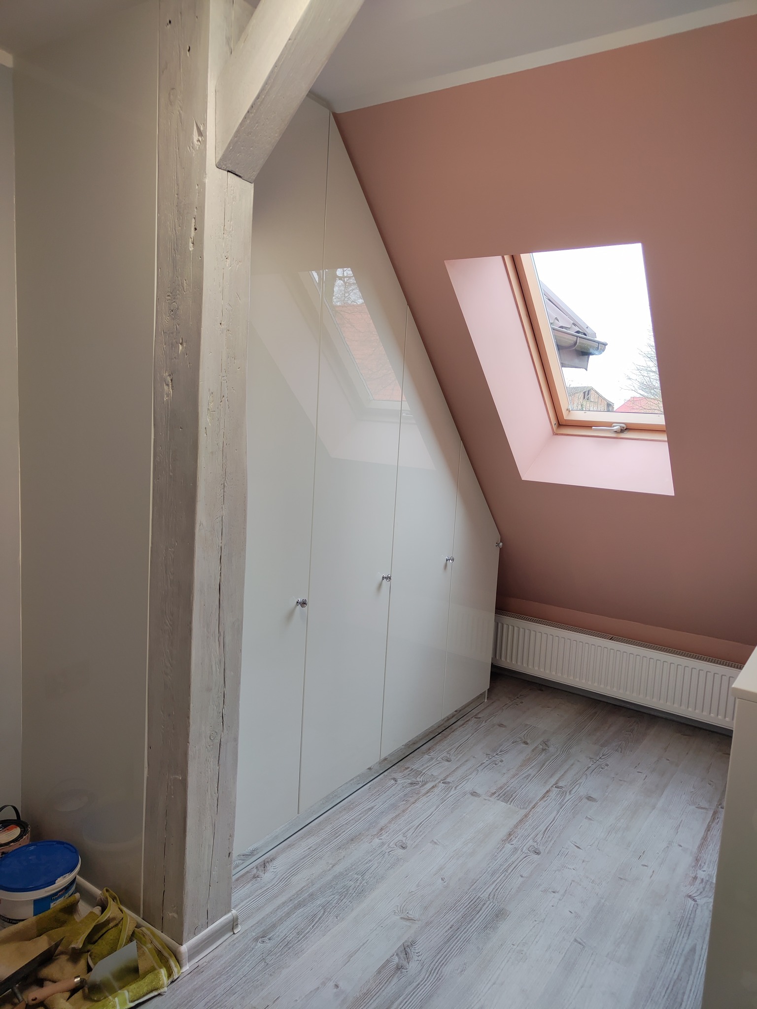 Biała szafa wnękowa na poddaszu, z drewnianą belką i oknem dachowym. Widoczna podłoga z paneli i fragment grzejnika. U dołu narzędzia i farby.