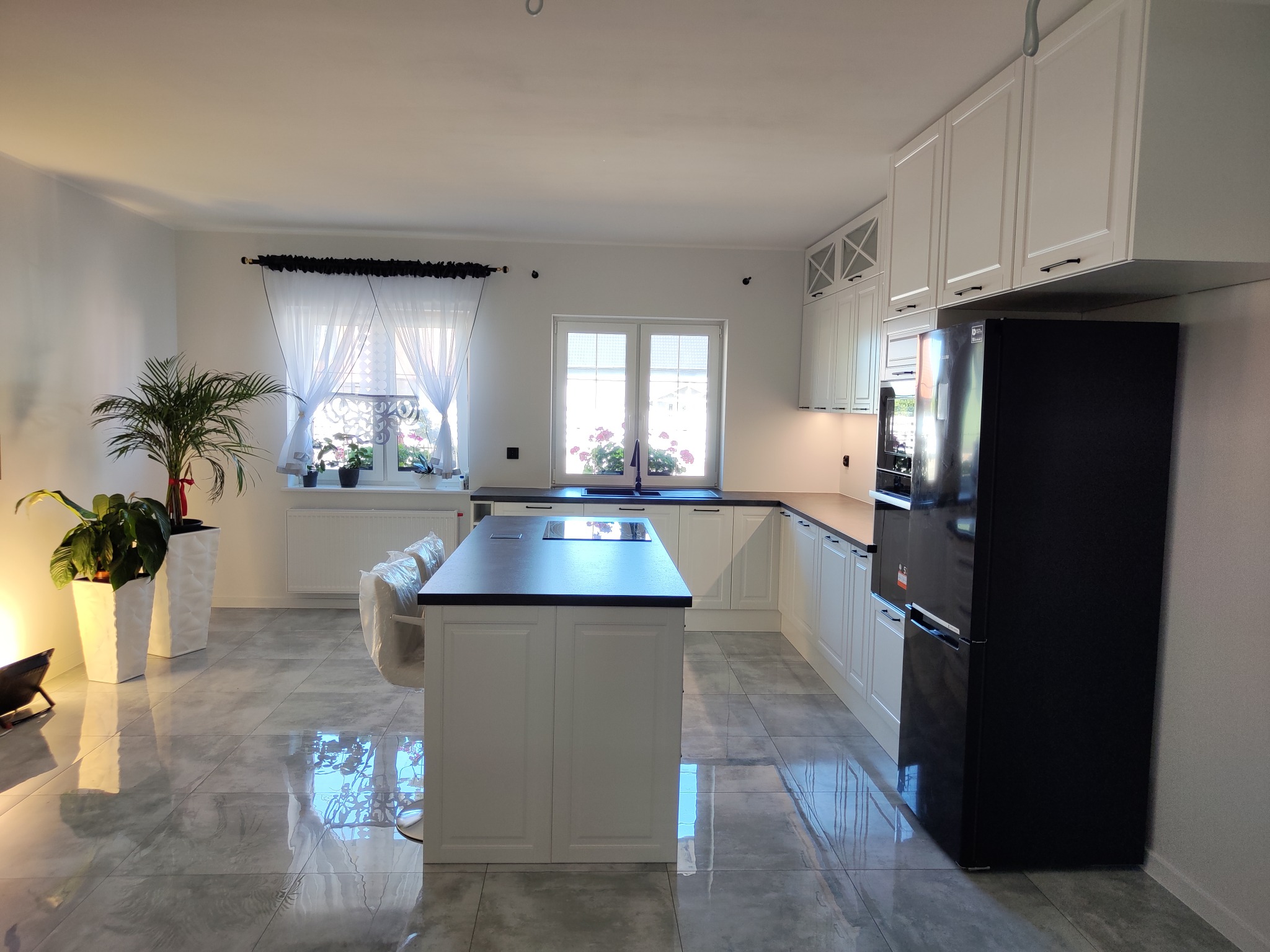 Jasna kuchnia z wyspą i czarną lodówką. Białe szafki z ciemnymi uchwytami, szare płytki podłogowe z połyskiem, rośliny doniczkowe w białych donicach.