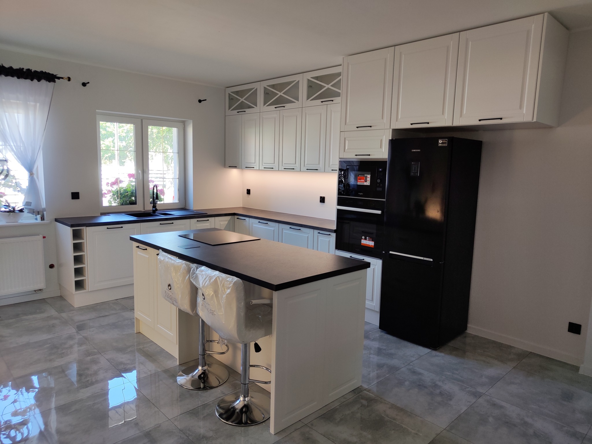 Jasna kuchnia z wyspą i czarnymi akcentami: blaty, lodówka i okucia. Krzesła barowe w folii ochronnej. Nowoczesny design z frezowanymi frontami i przeszklonymi szafkami.