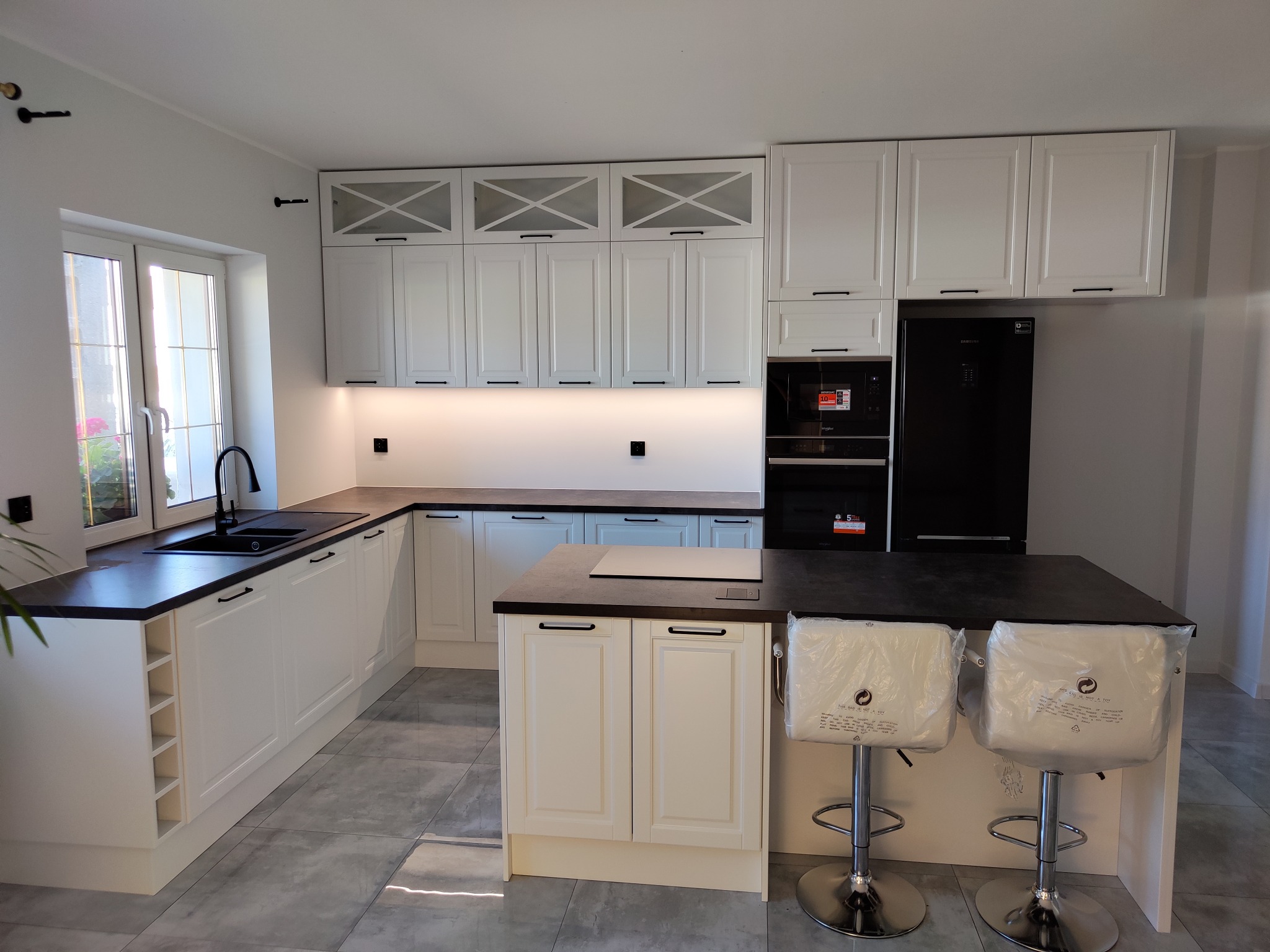 Jasna kuchnia z wyspą i czarnymi akcentami. Białe szafki z frezowanymi frontami, czarny blat i sprzęt AGD. Dwa krzesła barowe z folią ochronną.